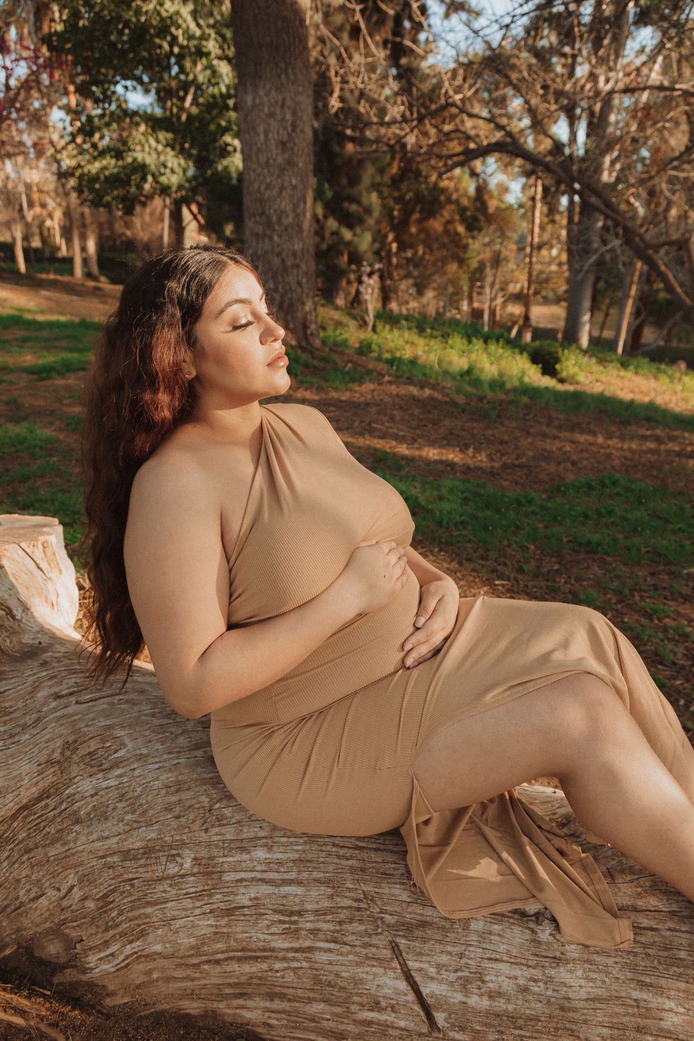 A woman in a beige dress poses on a fallen log in a sunlit forest setting during golden hour.