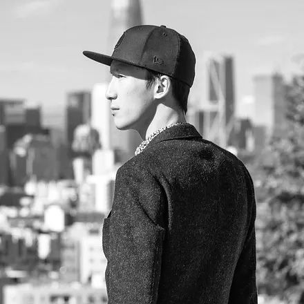 Young man in black snapback cap and chain necklace poses with city skyline in black and white photo.