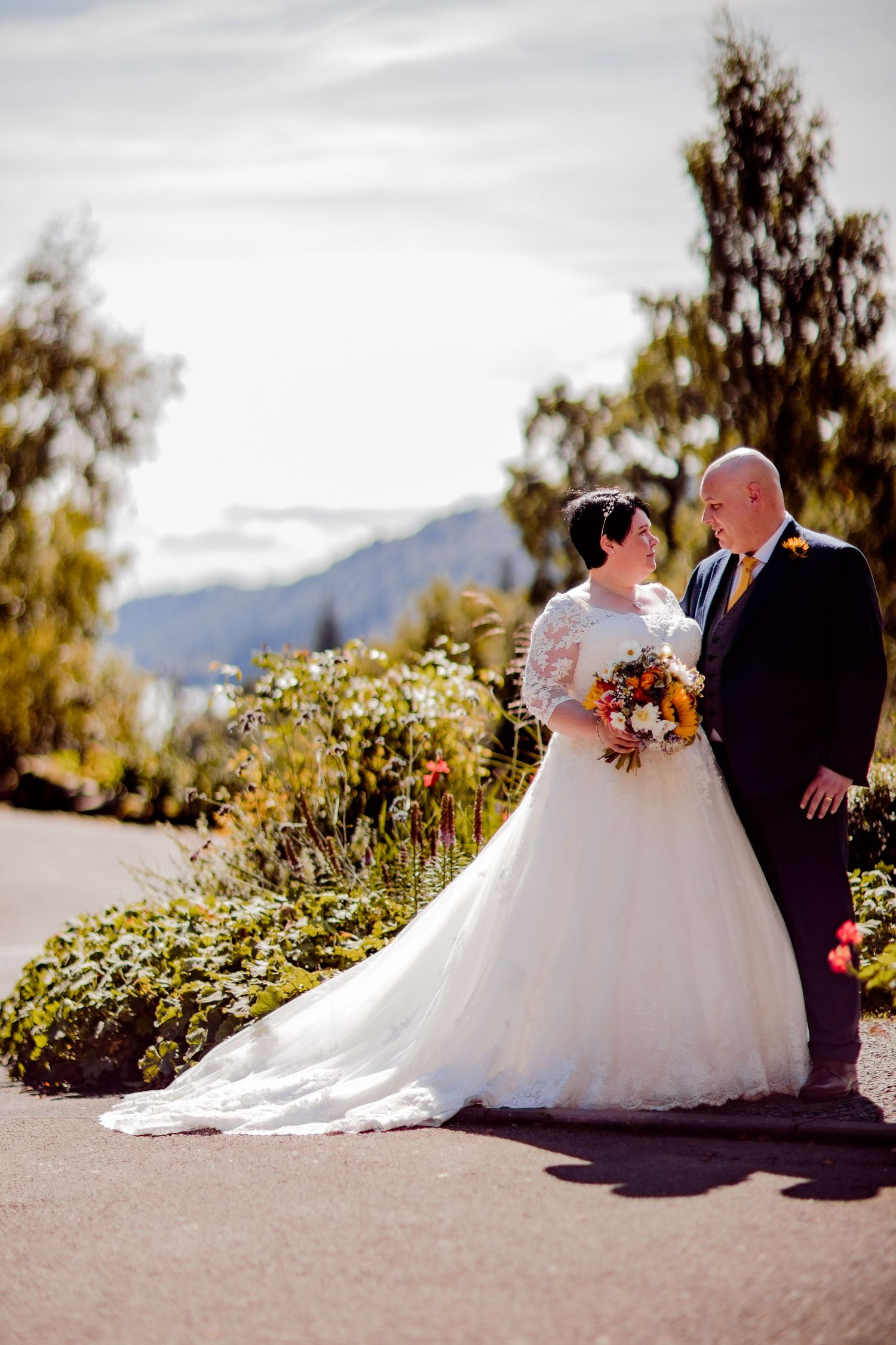 A wedding couple posing on a path with mountain scenery and colorful flowers in the background.
