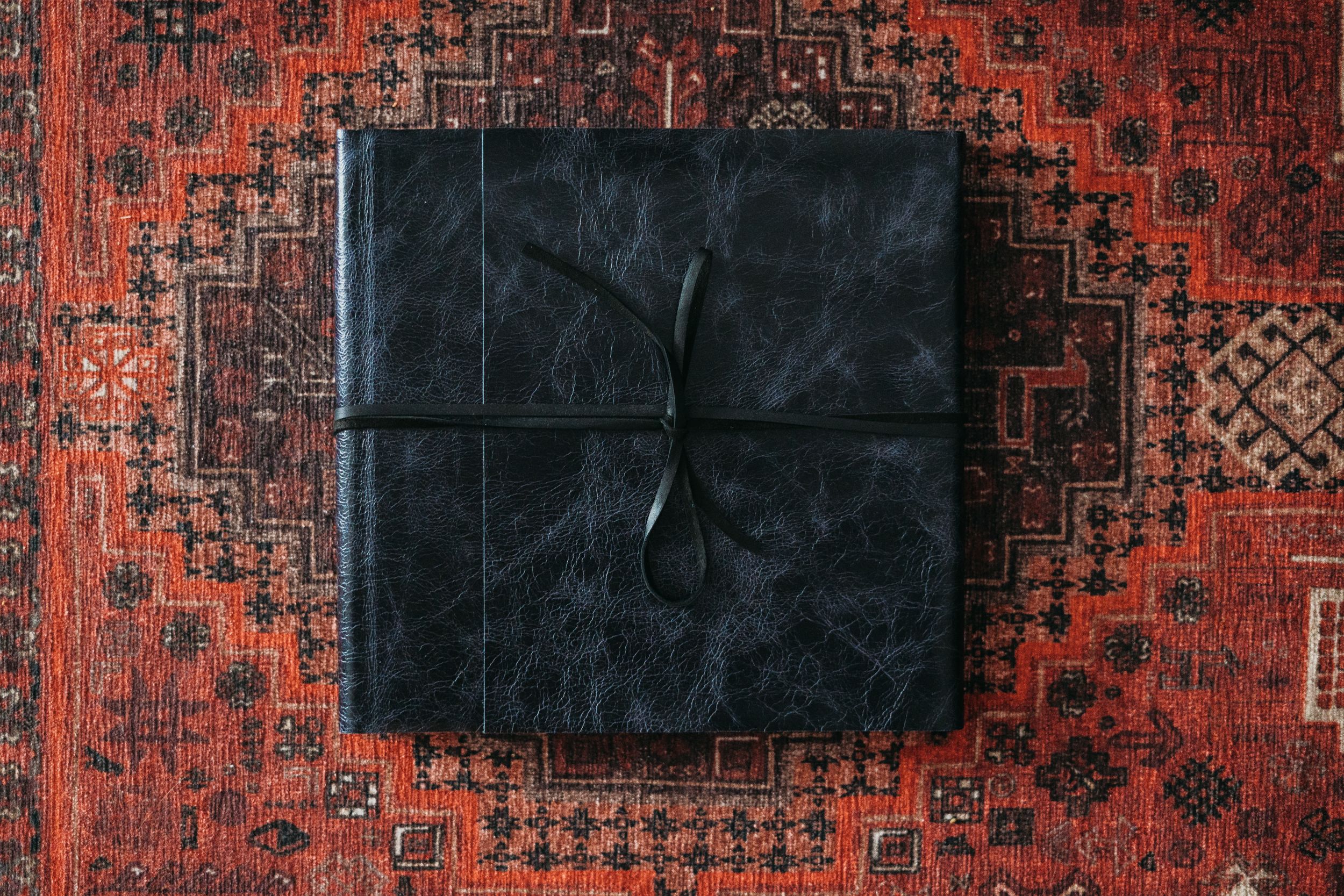 Square black leather album centered on ornate red patterned rug.