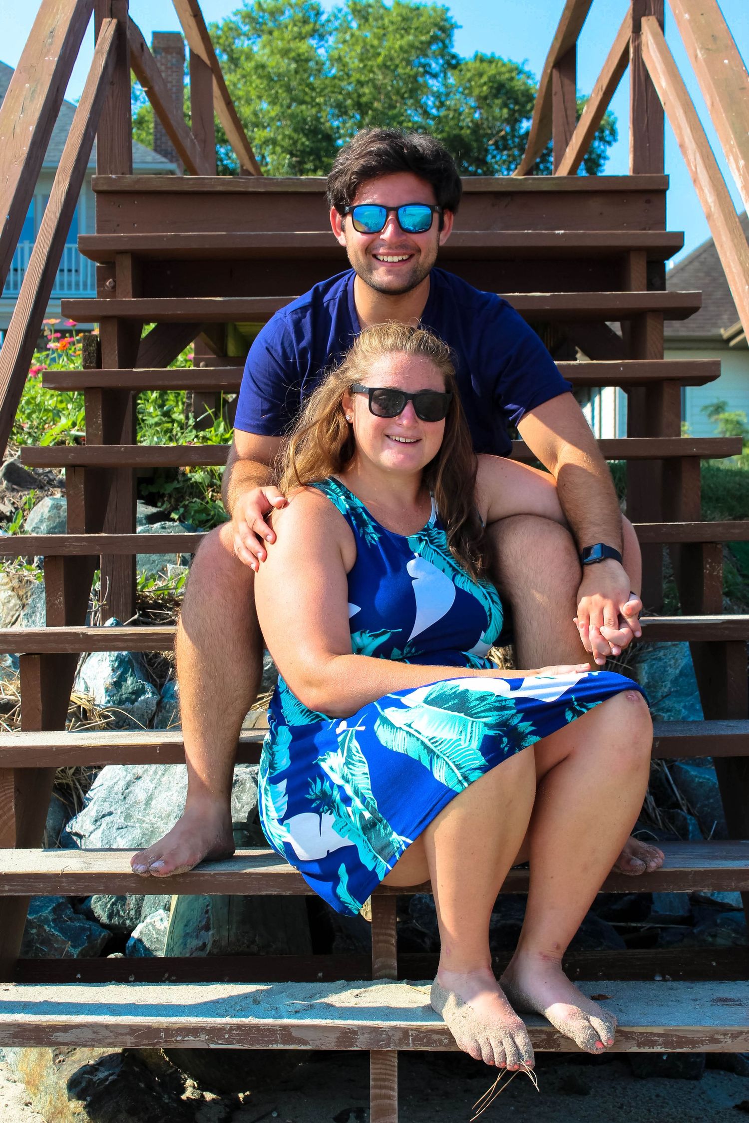 A couple wearing sunglasses sits together on wooden beach stairs on a sunny day.