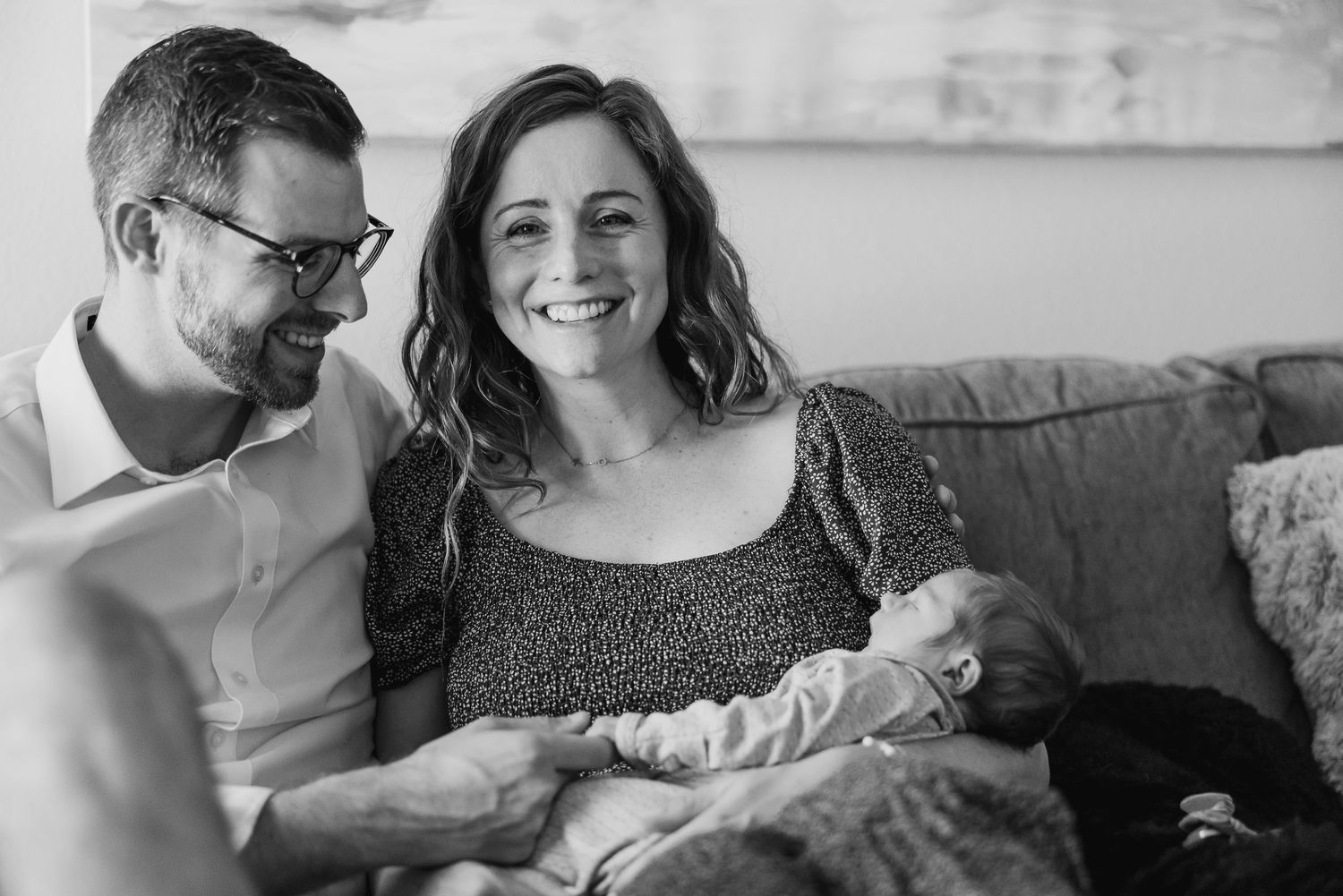 Candid black and white photo of new parents cuddling their tiny baby on a couch.