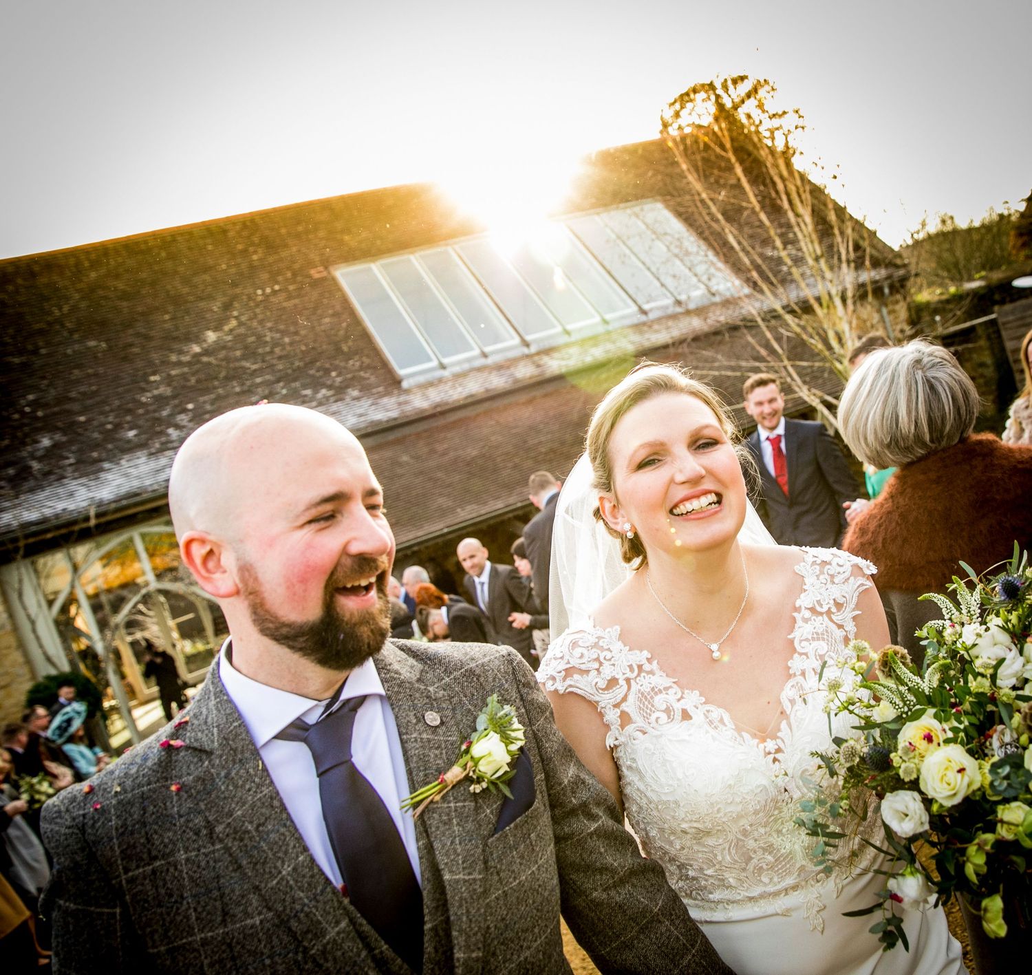 Sun-flared portrait of happy newlyweds with guests in background at outdoor venue.