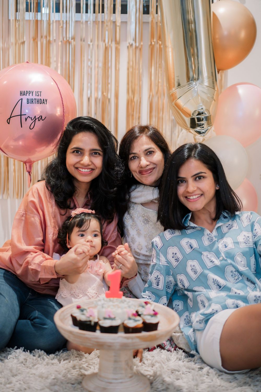 First birthday celebration with pink and gold party decorations and cupcakes on white carpet.