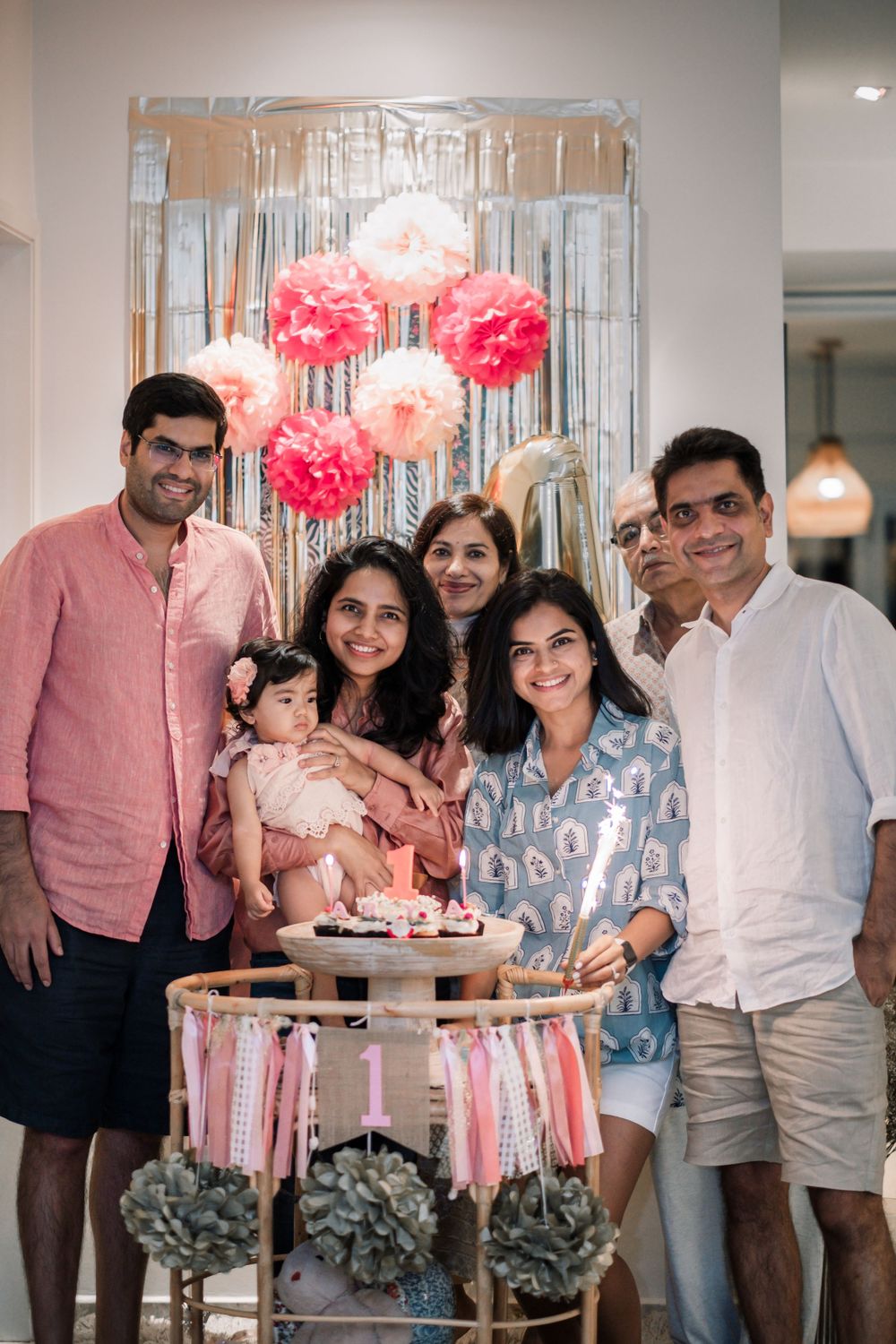 Family gathered around first birthday decorations with pink pom poms and metallic streamers.