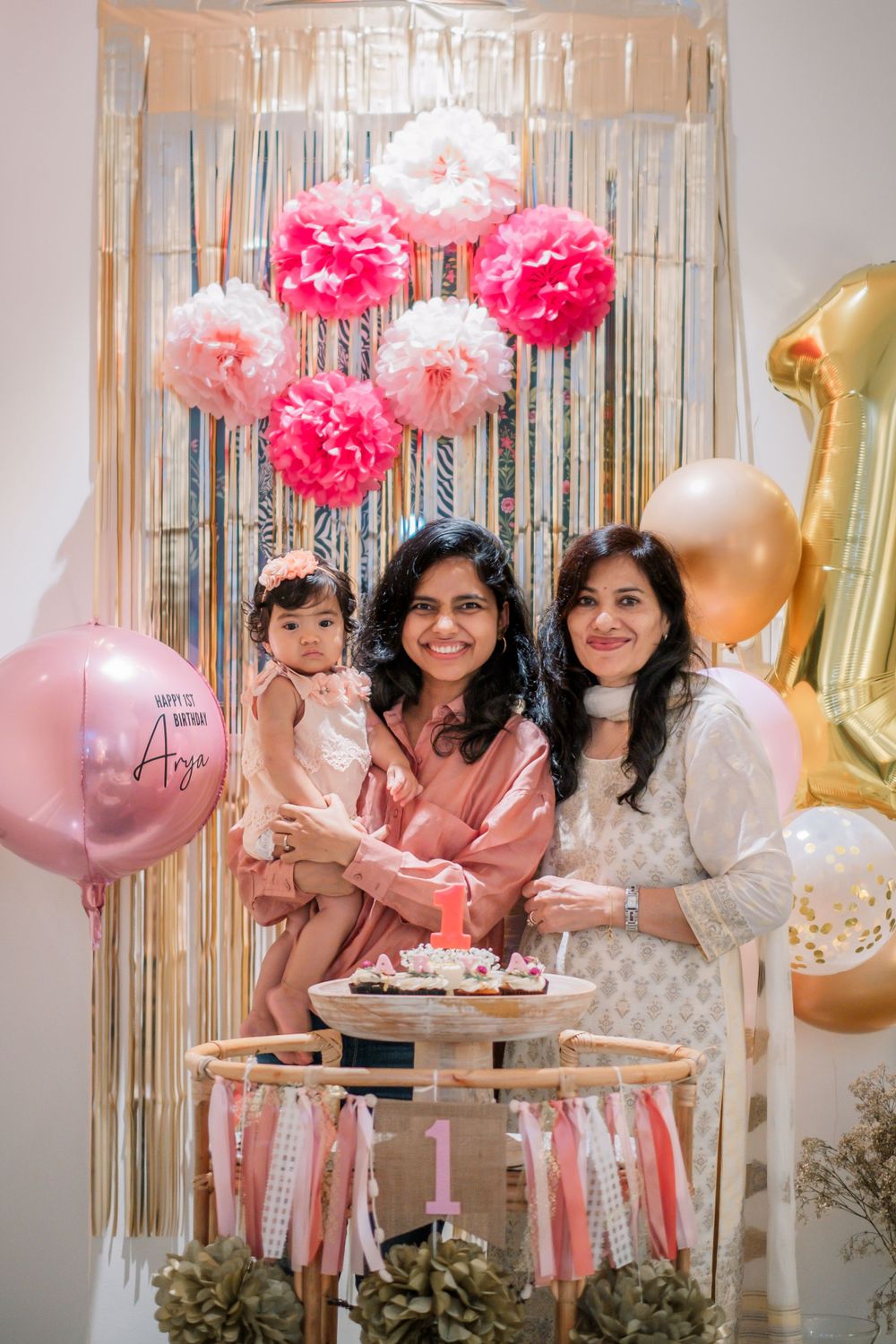 Pink and gold first birthday party decor with tissue pom poms and metallic balloons against a sparkly backdrop.