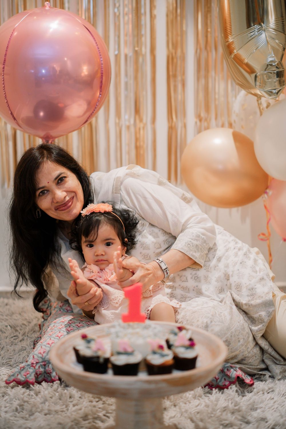 First birthday celebration with pink balloons and cupcakes on a decorative cake stand.