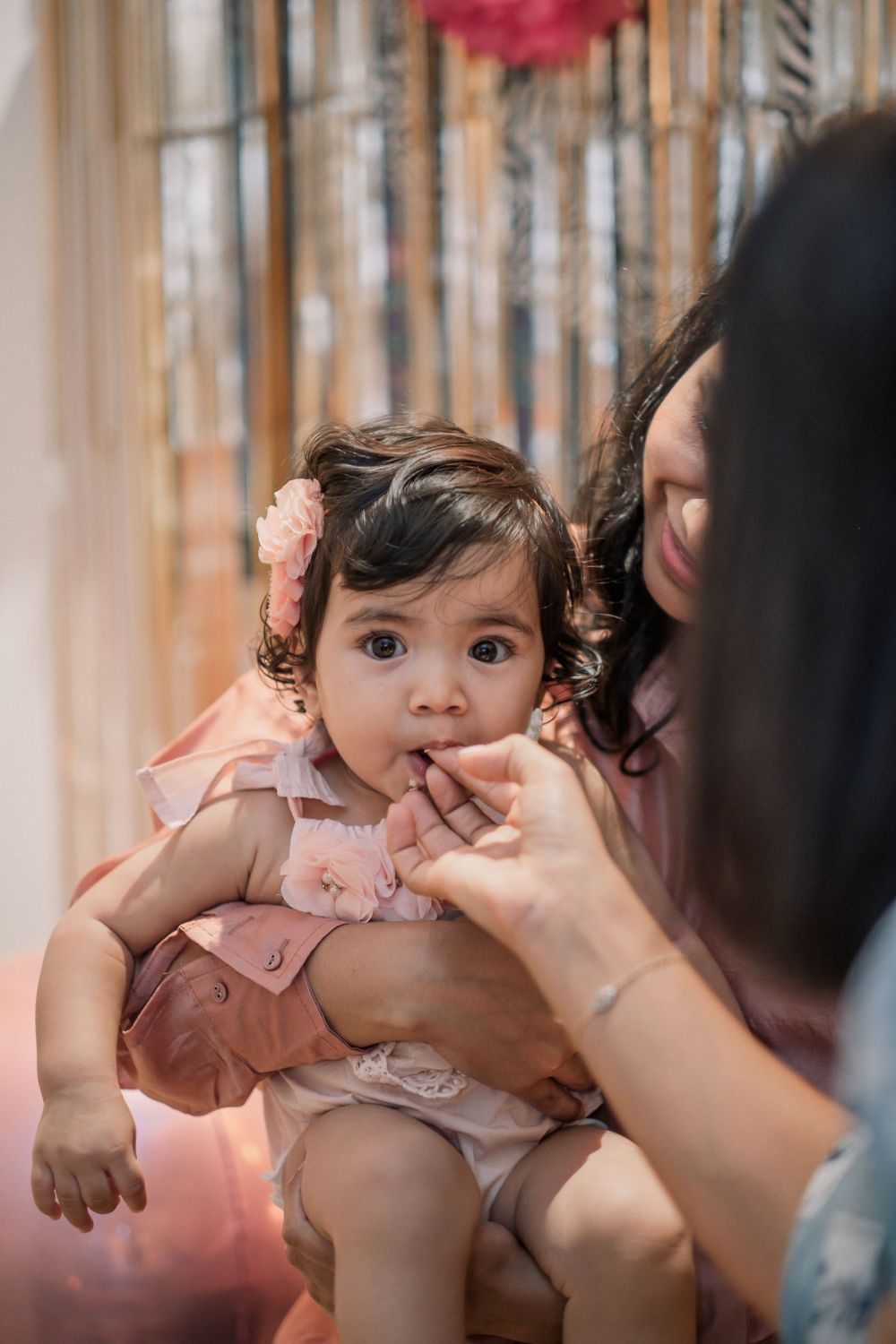 Baby in pink dress with curly hair being held at celebration with pink decorations.