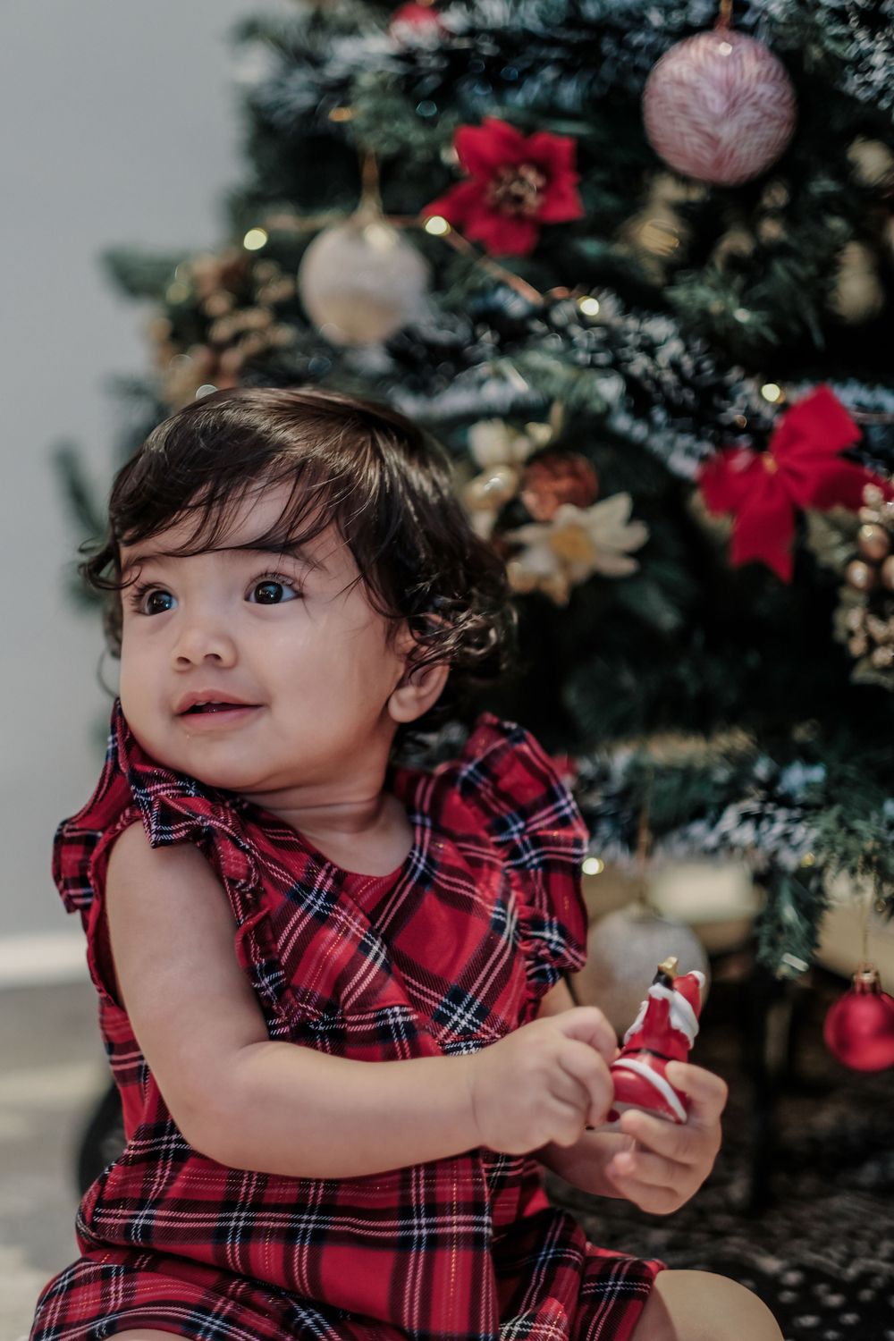 Toddler in plaid dress holding toy near decorated Christmas tree.