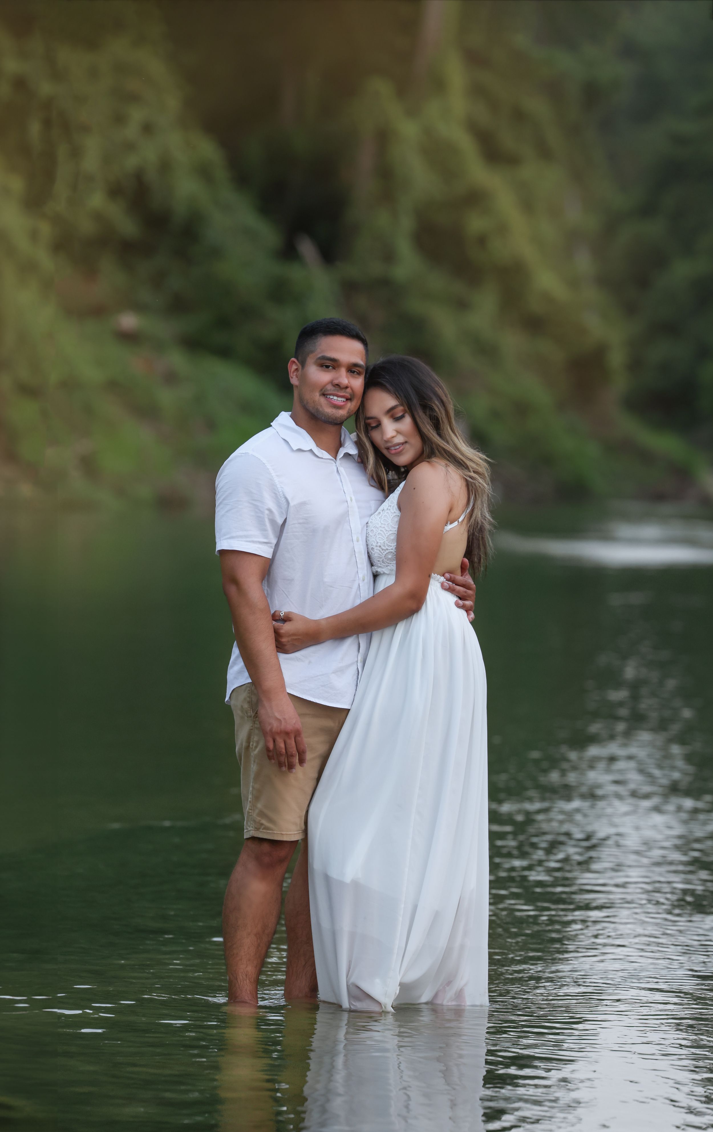 A romantic couple poses together in shallow water against a lush green backdrop.