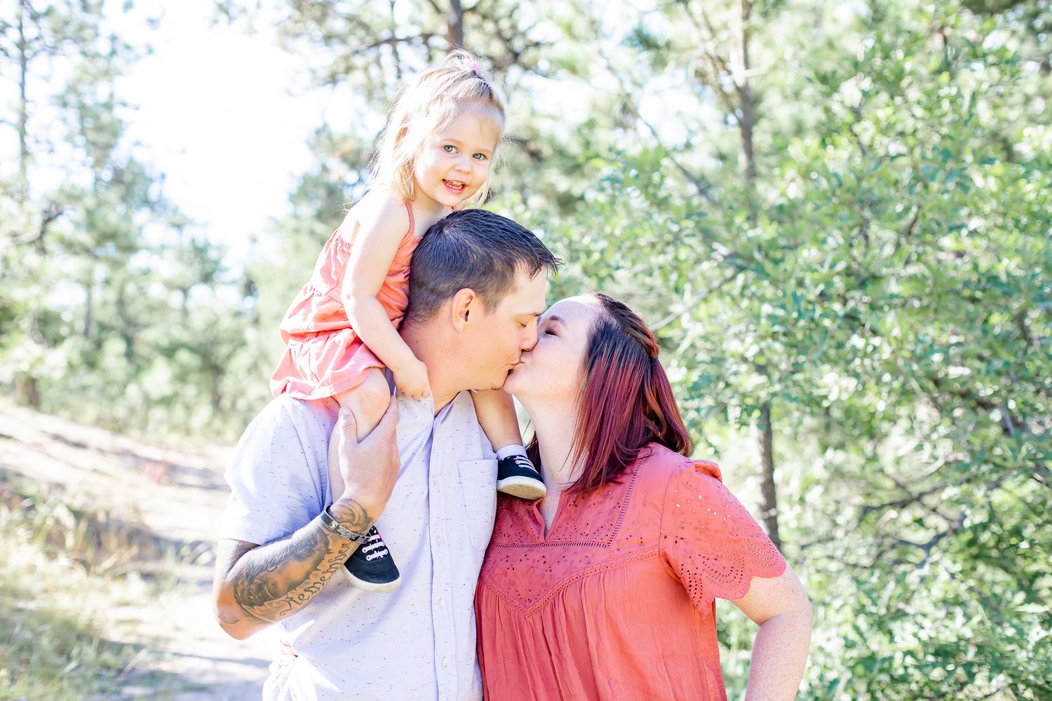 Mom and dad kiss while toddler daughter smiles from dad's shoulders in Black Forest, Colorado.