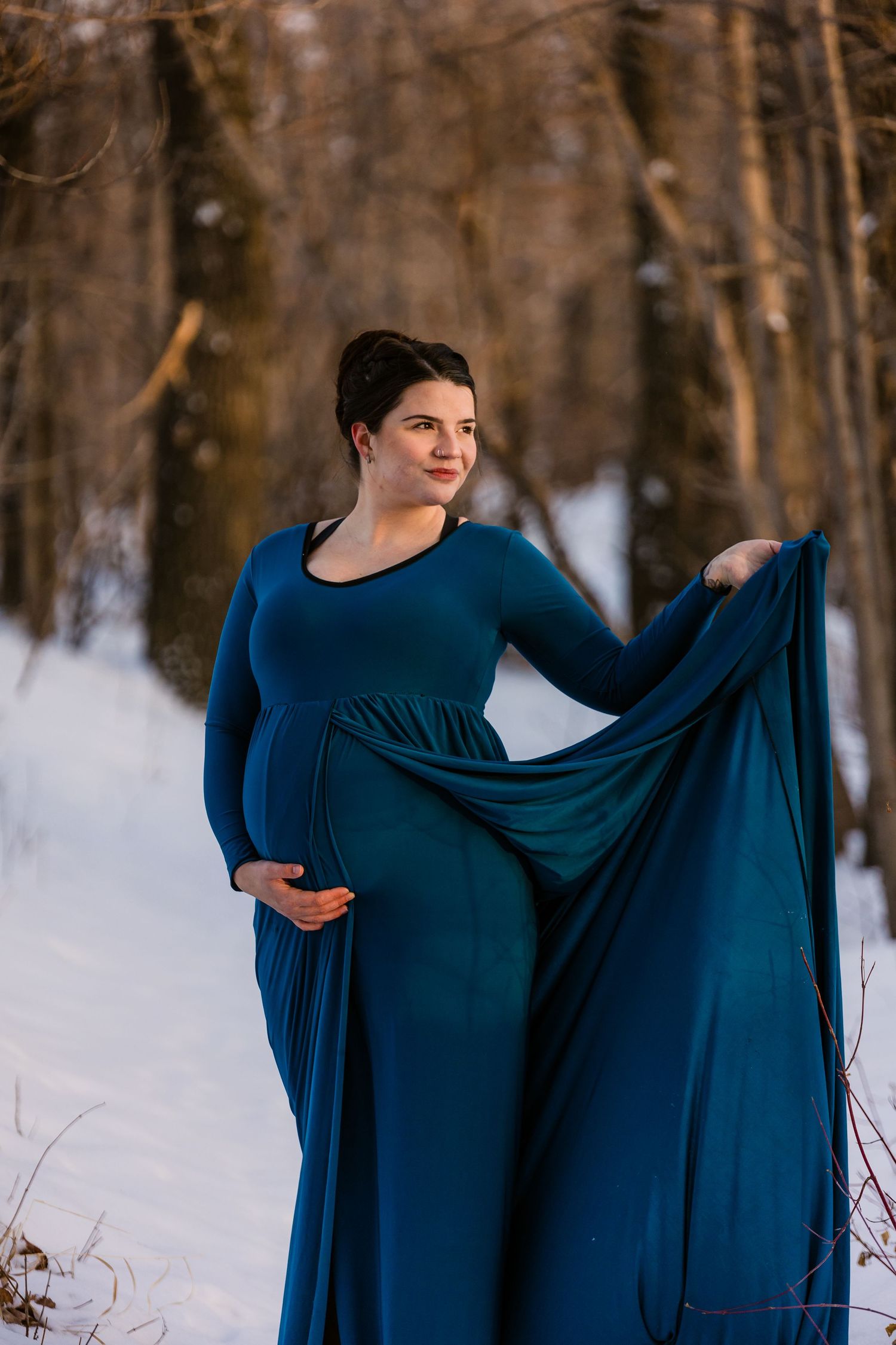 Maternity portrait in flowing teal gown against snowy winter forest backdrop.