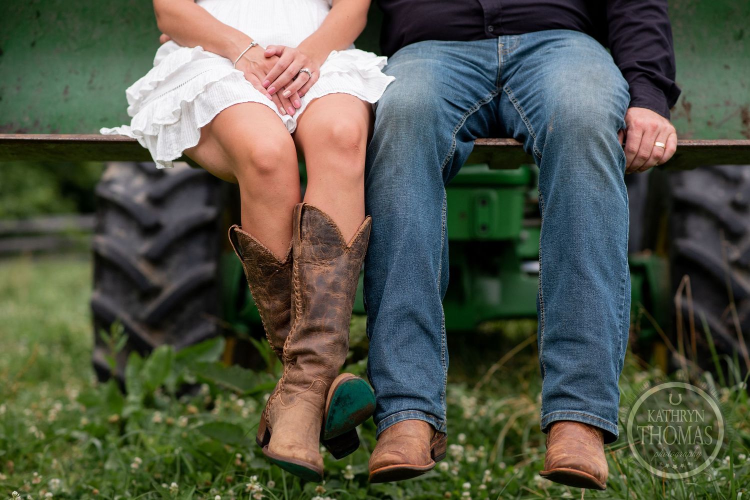 Two pairs of legs wearing jeans and cowboy boots sit on a green tractor in a rural setting.