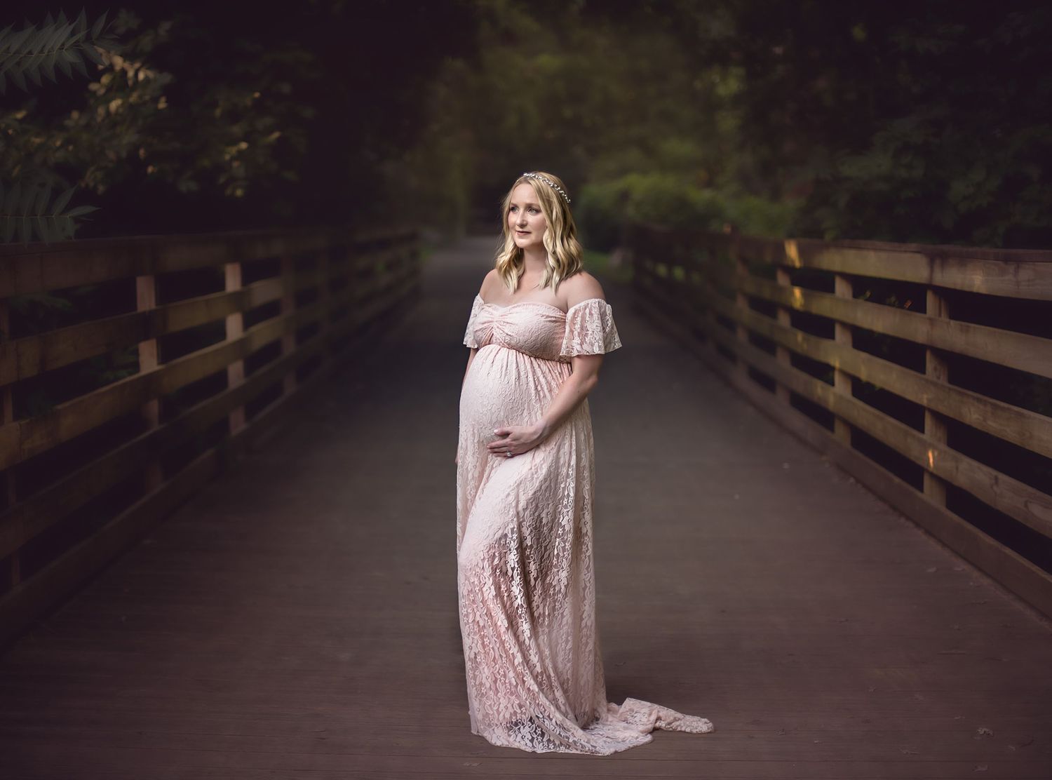 Woman in lace gown poses gracefully on wooden bridge surrounded by trees at dusk.