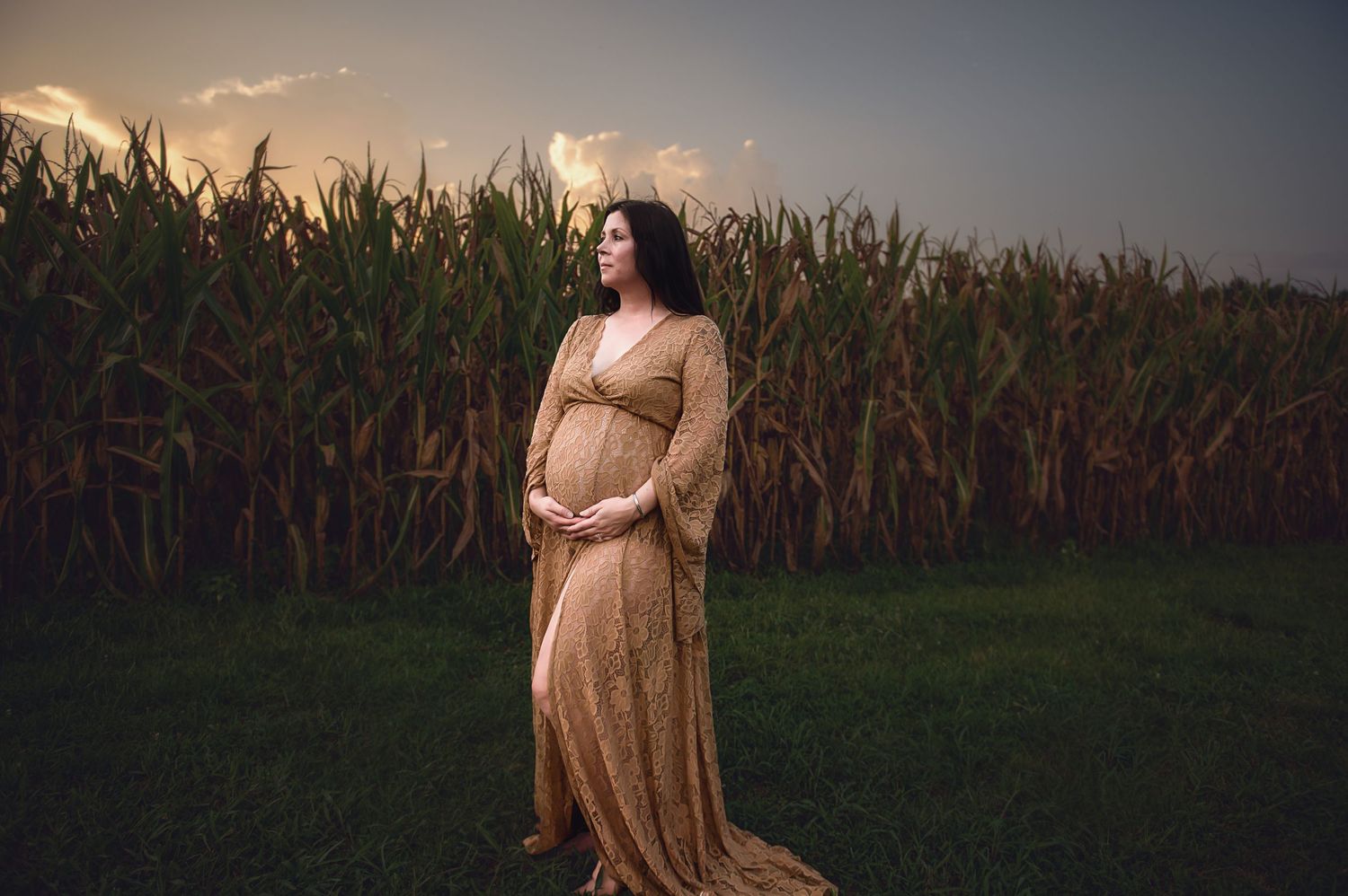 Woman in flowing beige dress stands elegantly in front of cornfield at sunset.