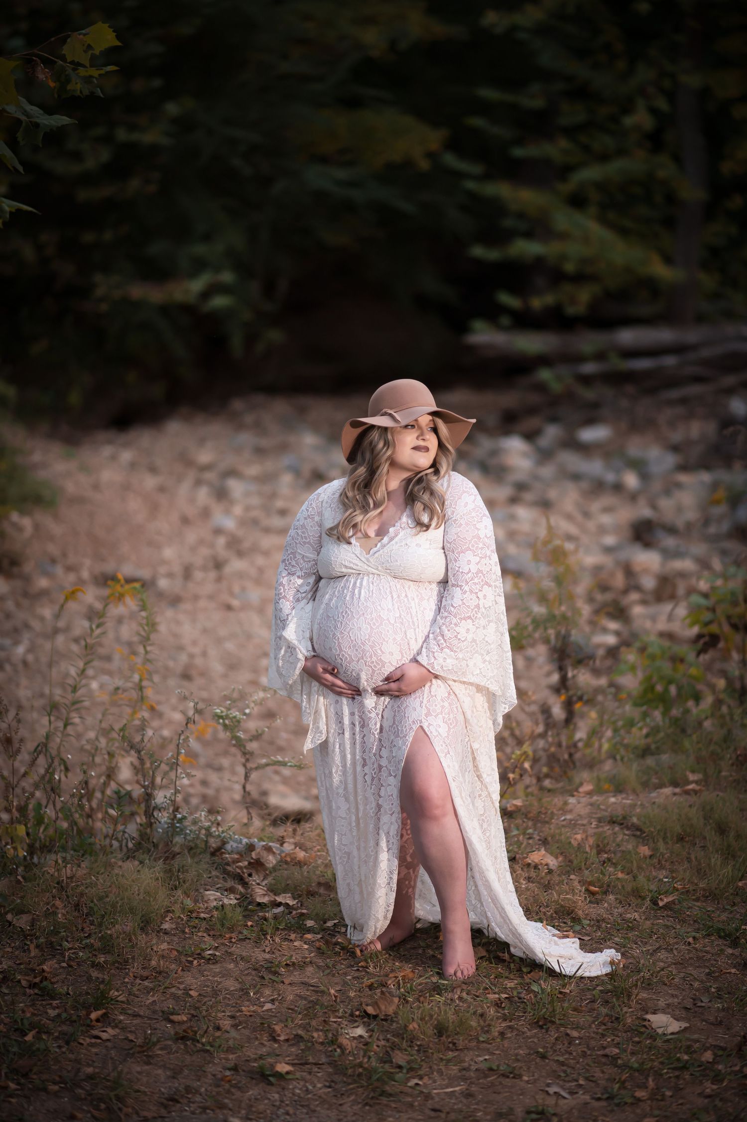 Bohemian maternity portrait in lace dress and floppy hat on a woodland trail.
