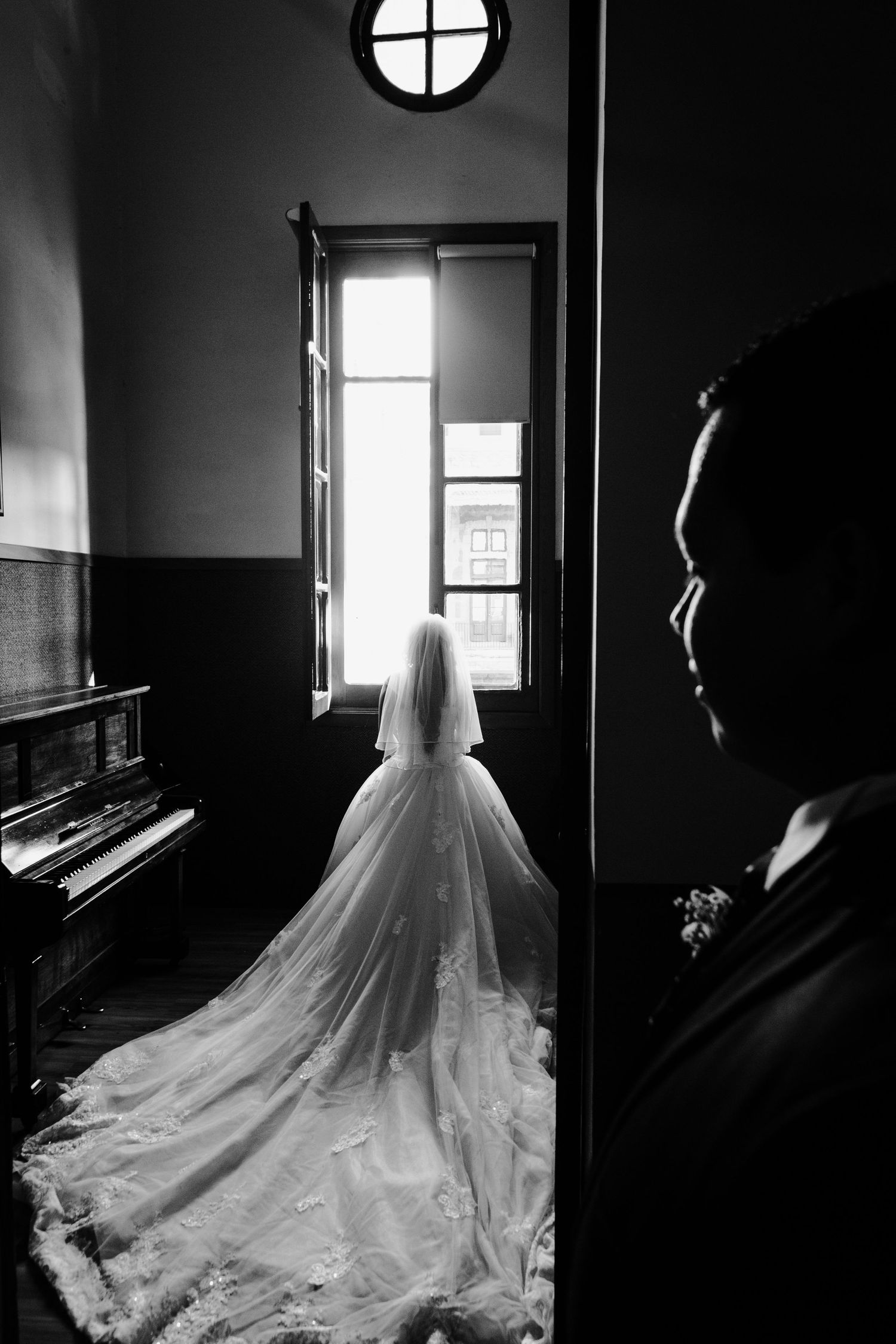 Dramatic black and white wedding photo with silhouette near piano and window.