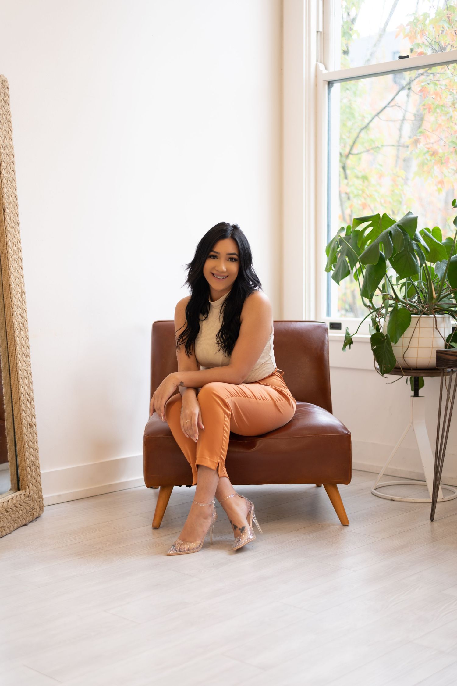 Hispanic woman sitting in studio on brown chair looking and smiling at camera.