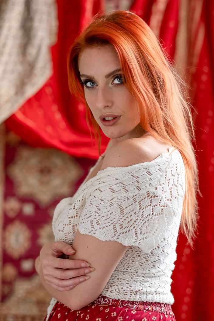 Portrait of a woman with red hair in white lace against red drapery background.
