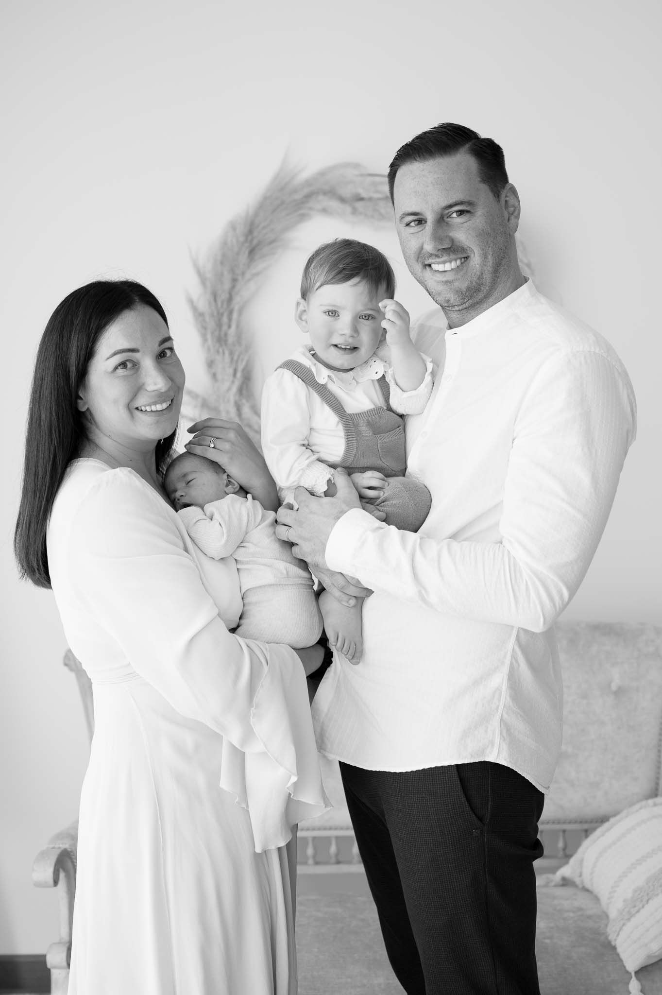 Family portrait in white showing parents holding two young children against a light background.