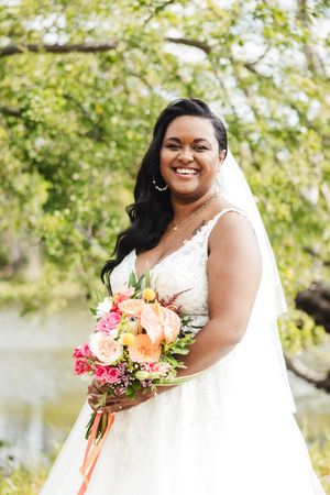 Bride in white gown holds vibrant pink and orange bouquet by waterside.