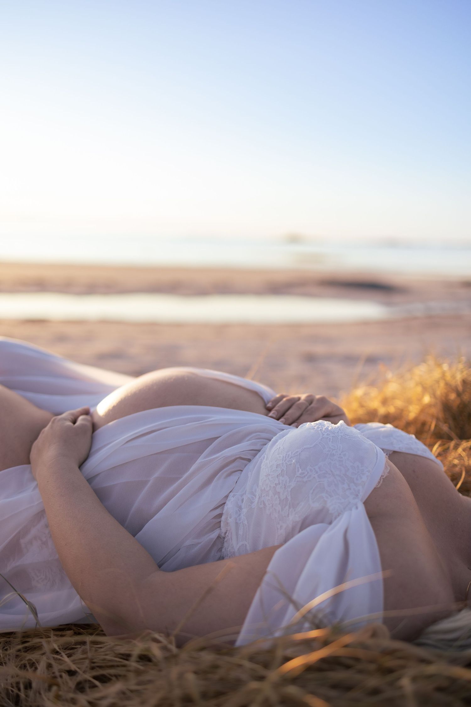 Dreamy maternity photo showing a figure in flowing white fabric resting on beach sand.