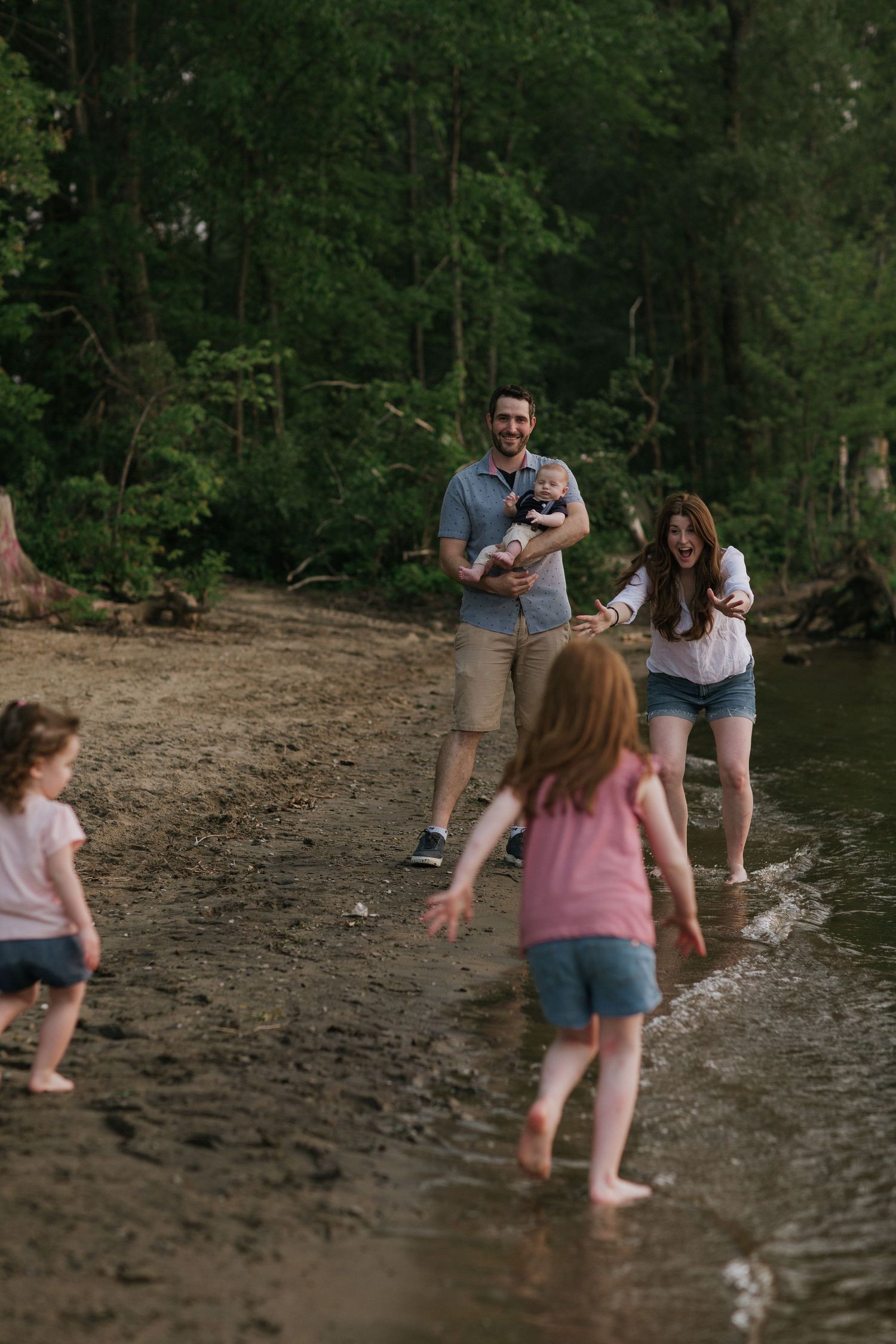 Children and adults splashing and playing in shallow water near a wooded shoreline.