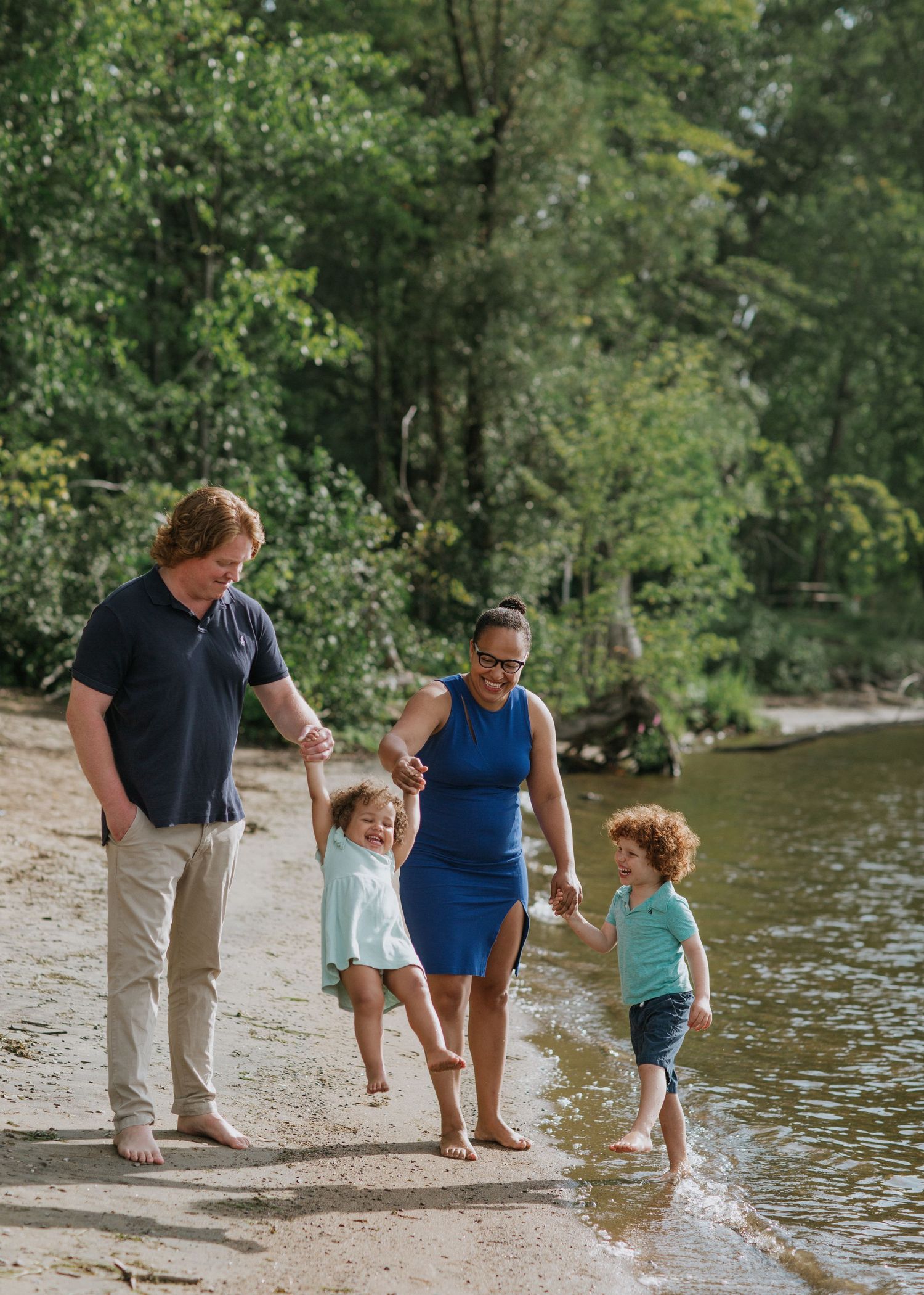 Family group walking along shoreline of lake surrounded by green trees.