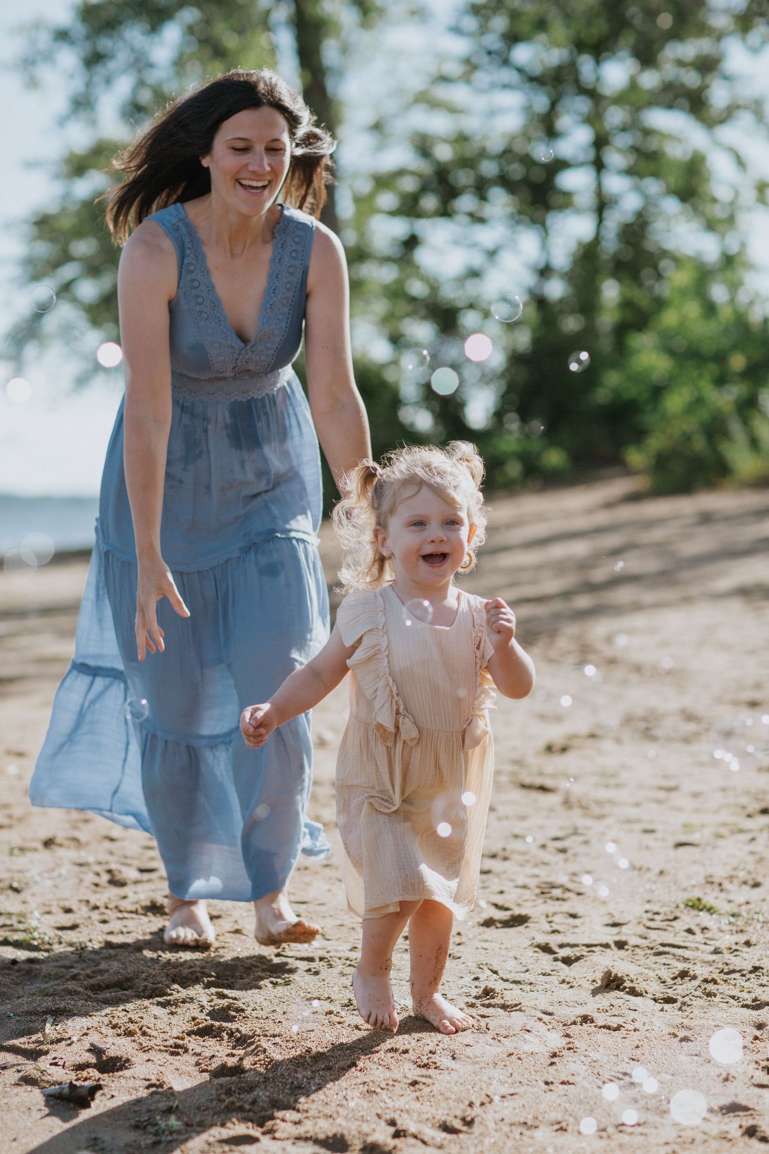 Joyful child in light colored clothing runs along beach path with companion in blue dress.