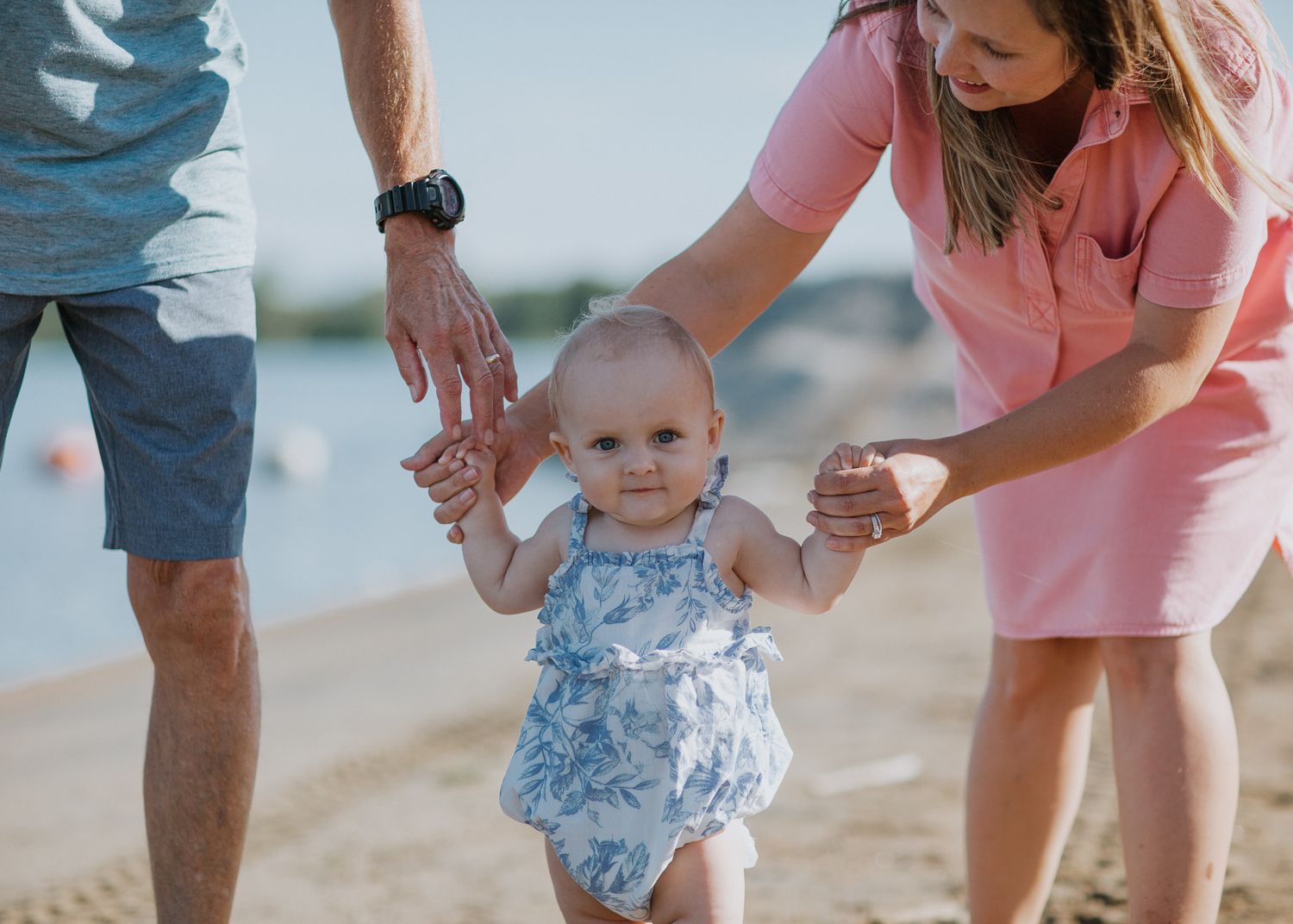 Parents helping toddler take first steps on sandy beach in summer.