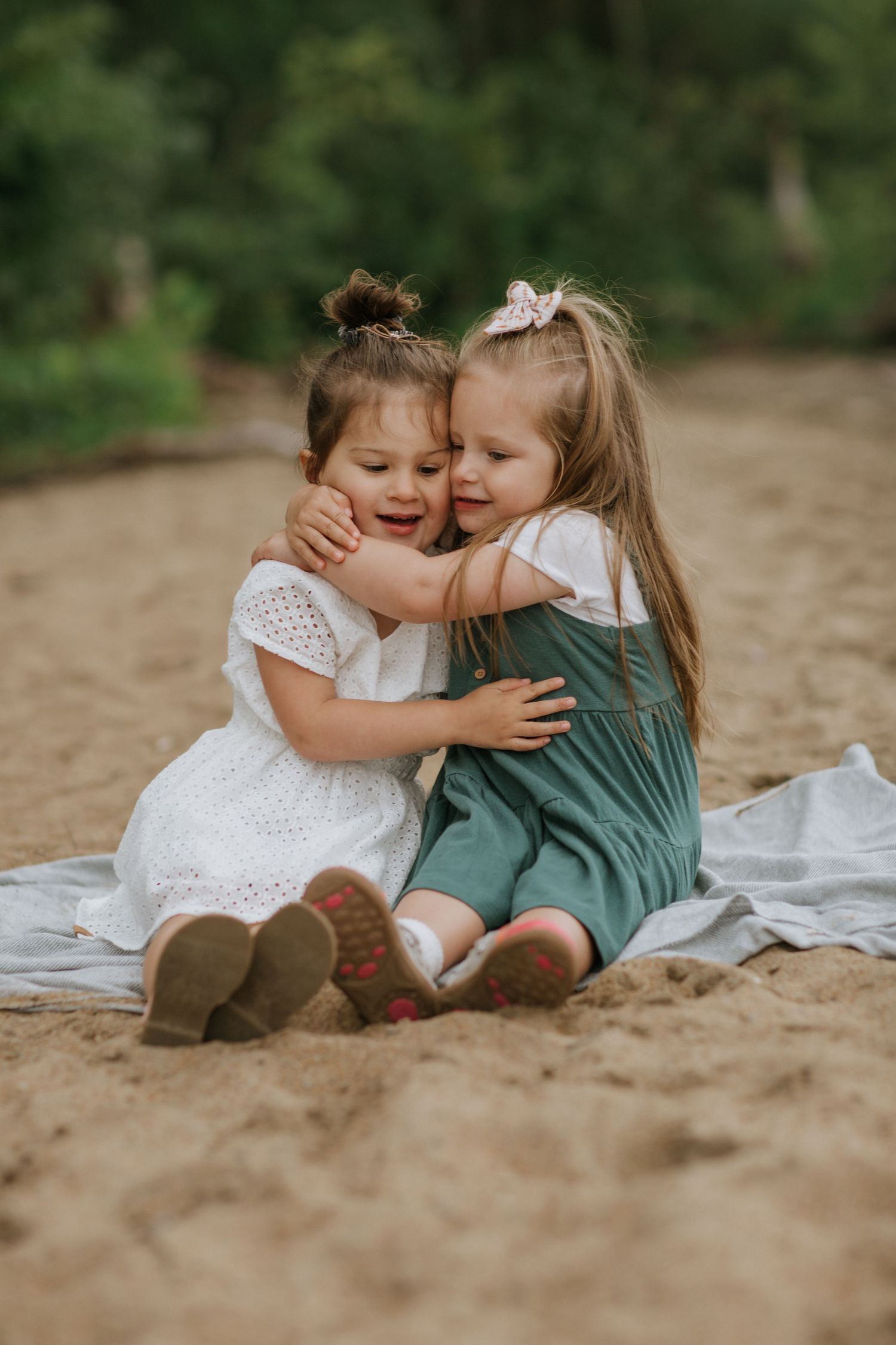 Two children in white and green dresses sharing a heartfelt hug on sandy ground.