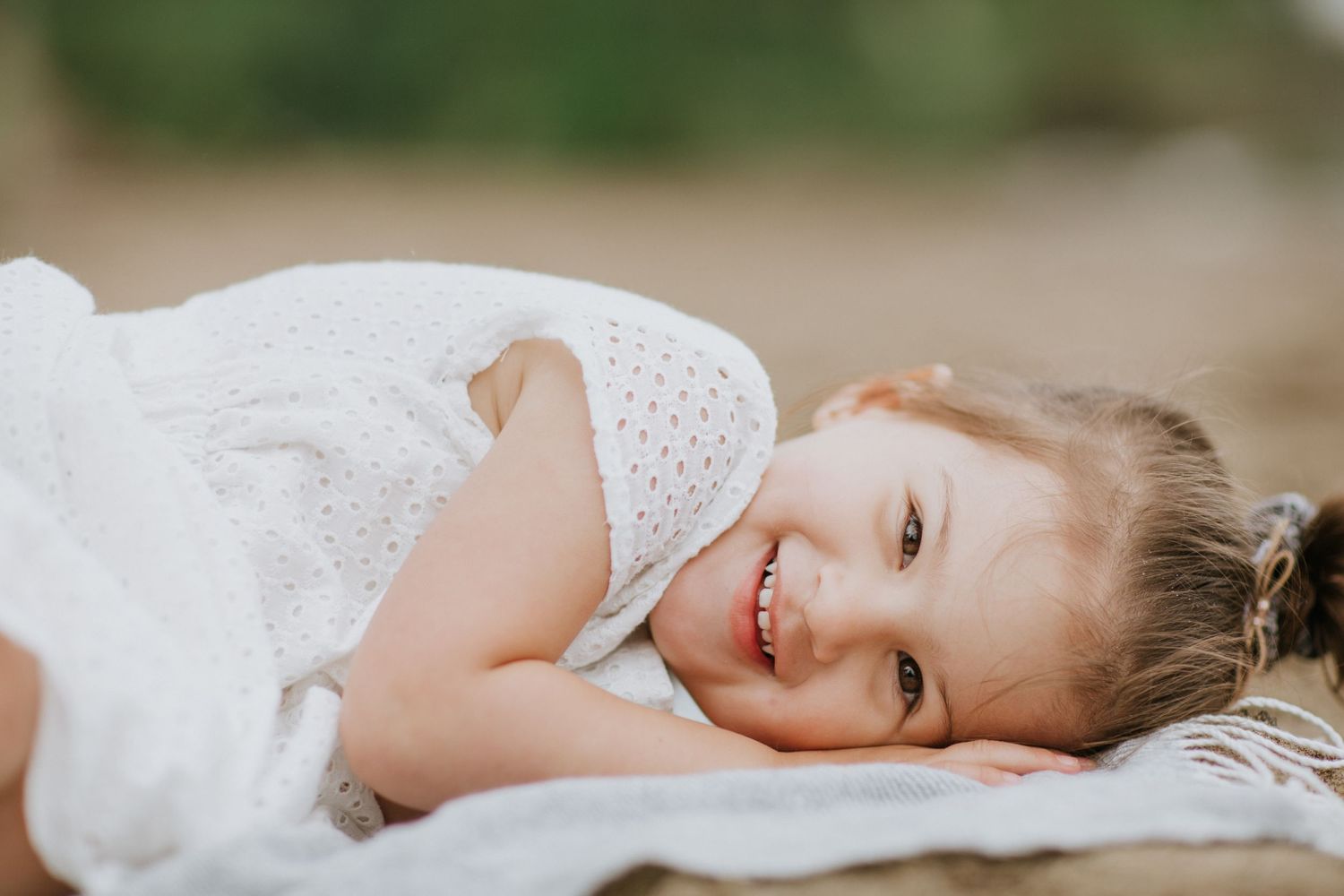 Young child in white lace dress lying on blanket outdoors with joyful expression.