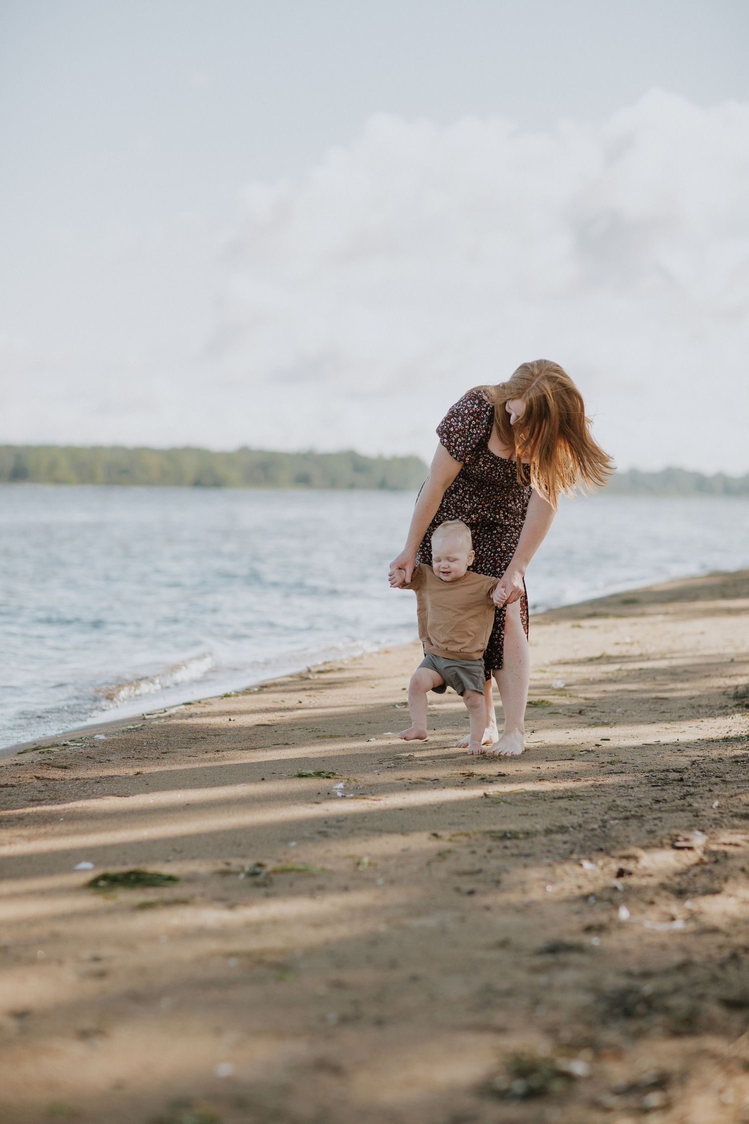 A person in a dress bends down to explore the sandy beach shoreline at sunset.