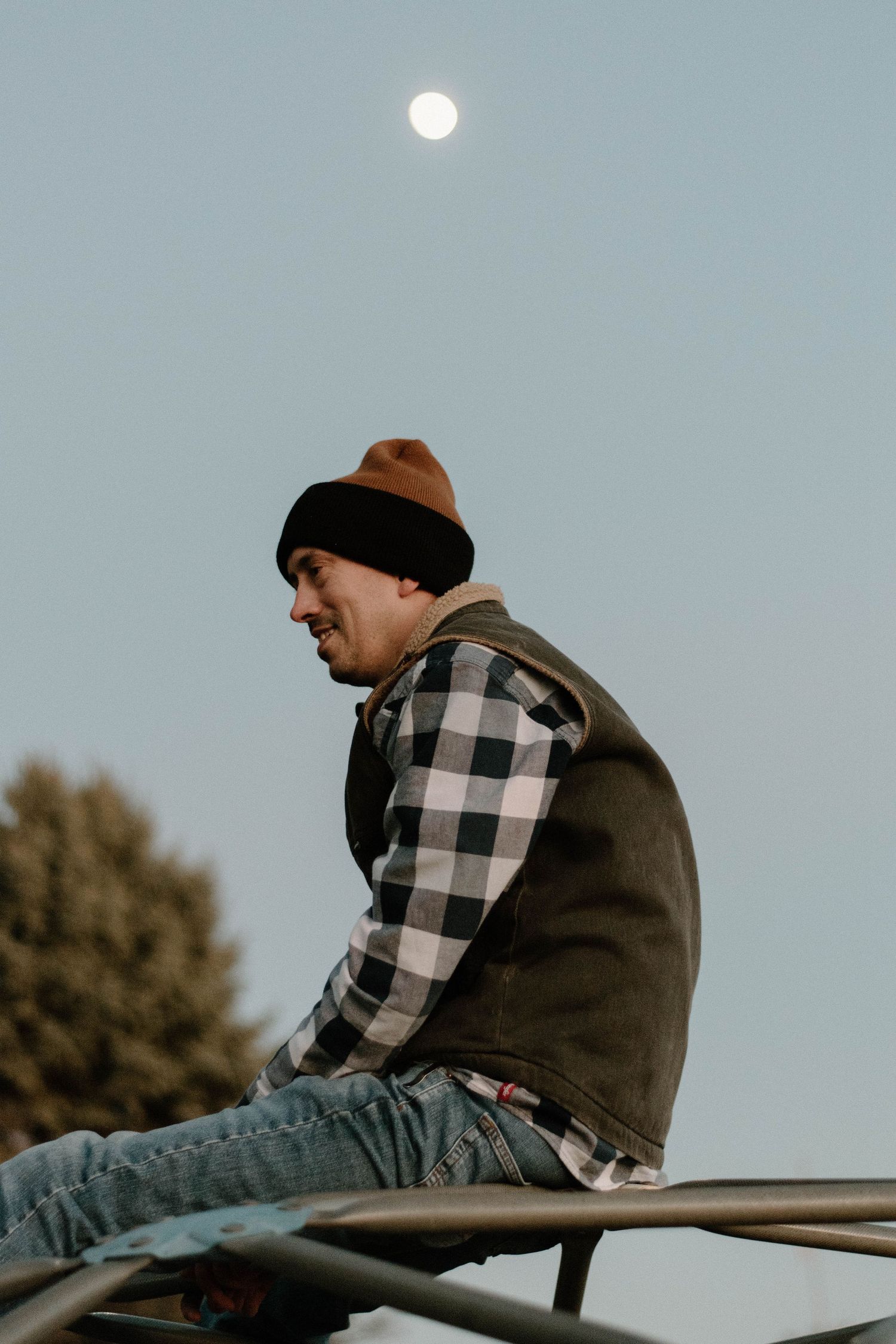 Person in plaid and beanie sits casually on metal structure with moon visible in sky.