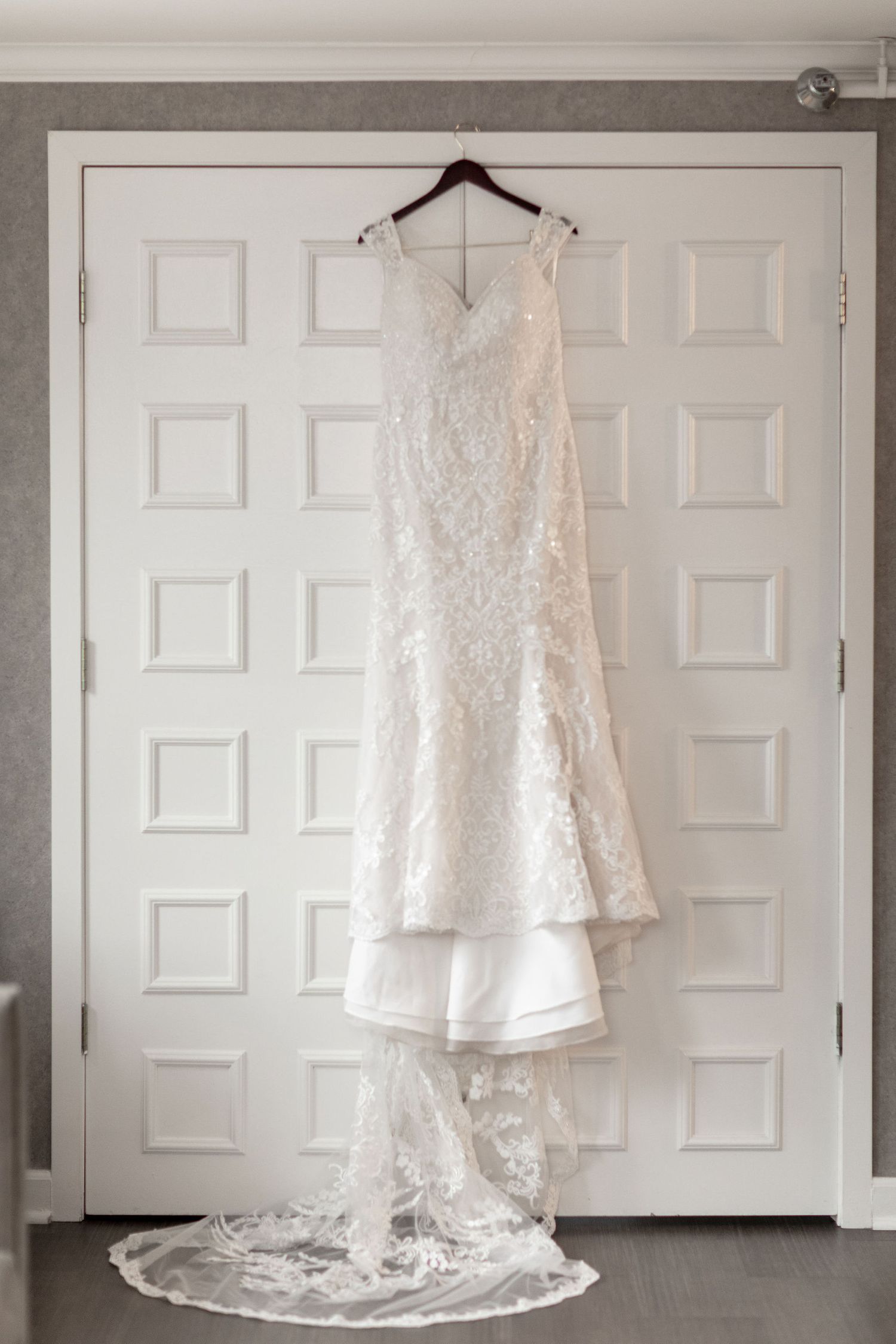 An elegant white lace wedding dress with a long train hanging on display against a white door.