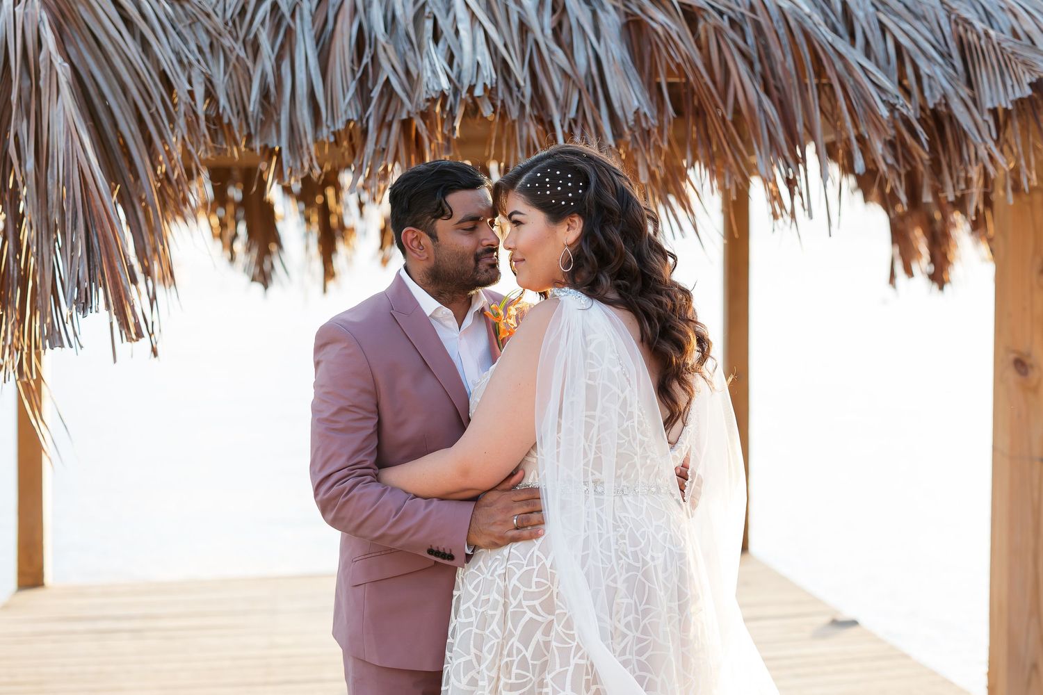 A couple embracing on a beach with a thatched roof structure in the background.