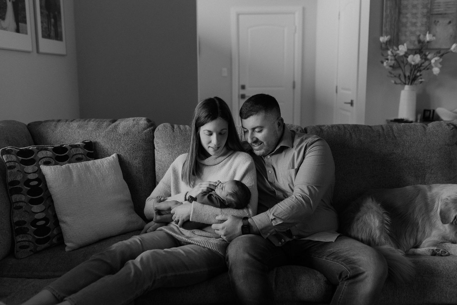 A tender family moment captured in black and white as parents cuddle with their newborn on a couch.