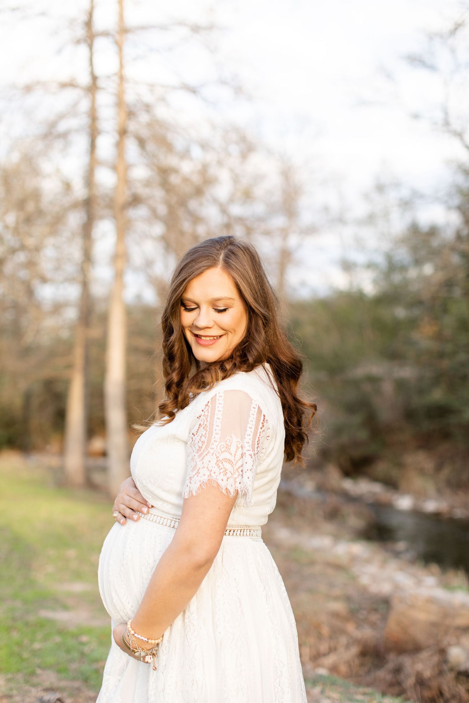 A pregnant woman in white lace dress smiles while posing by water.