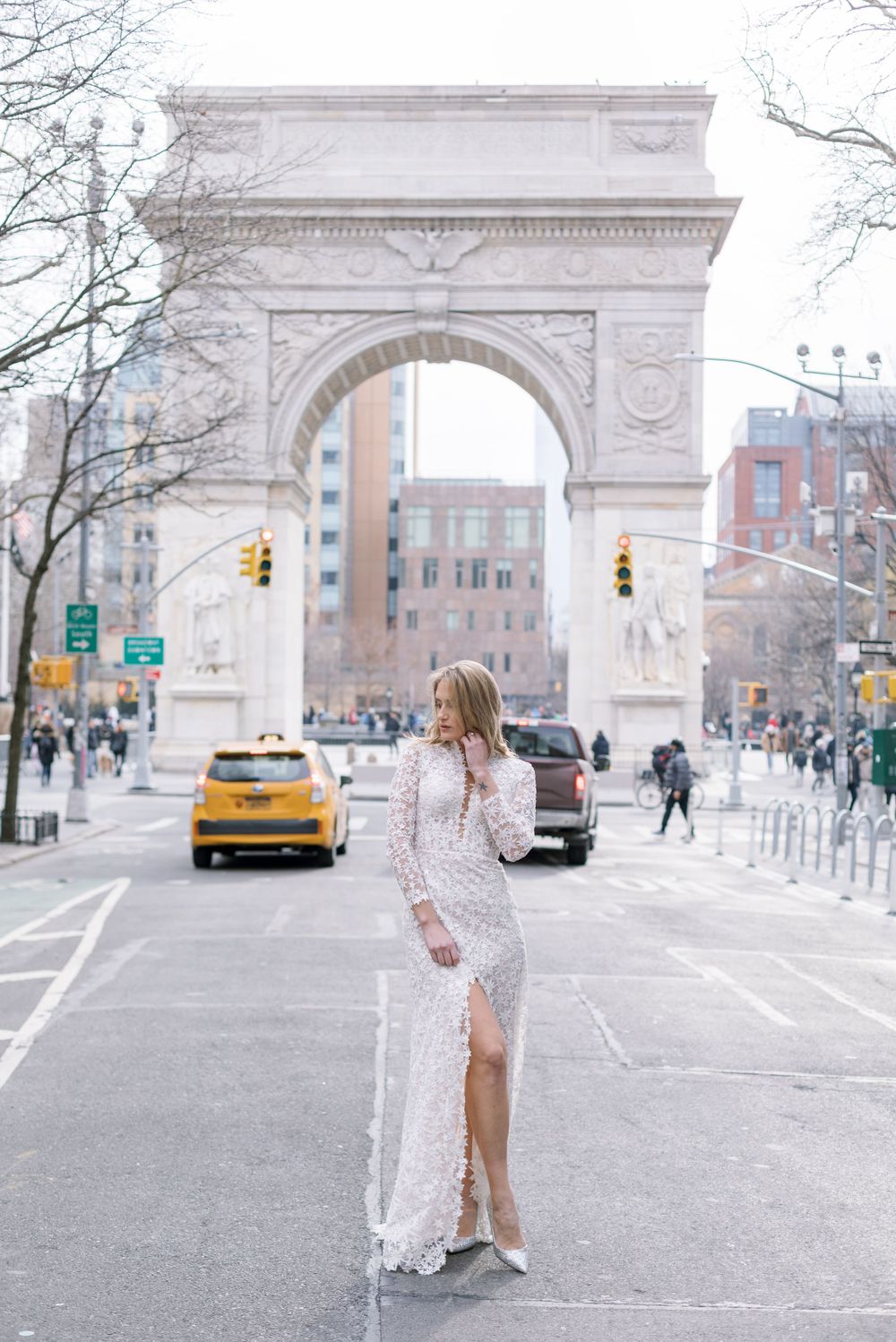 Bride in long sleeve lace gown poses in front of Washington Square Arch with NYC taxis passing by.