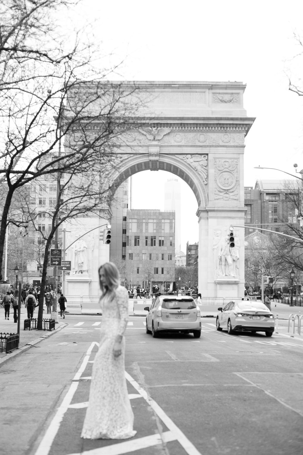 Black and white wide shot of bride in lace gown centered on empty street with historic arch.