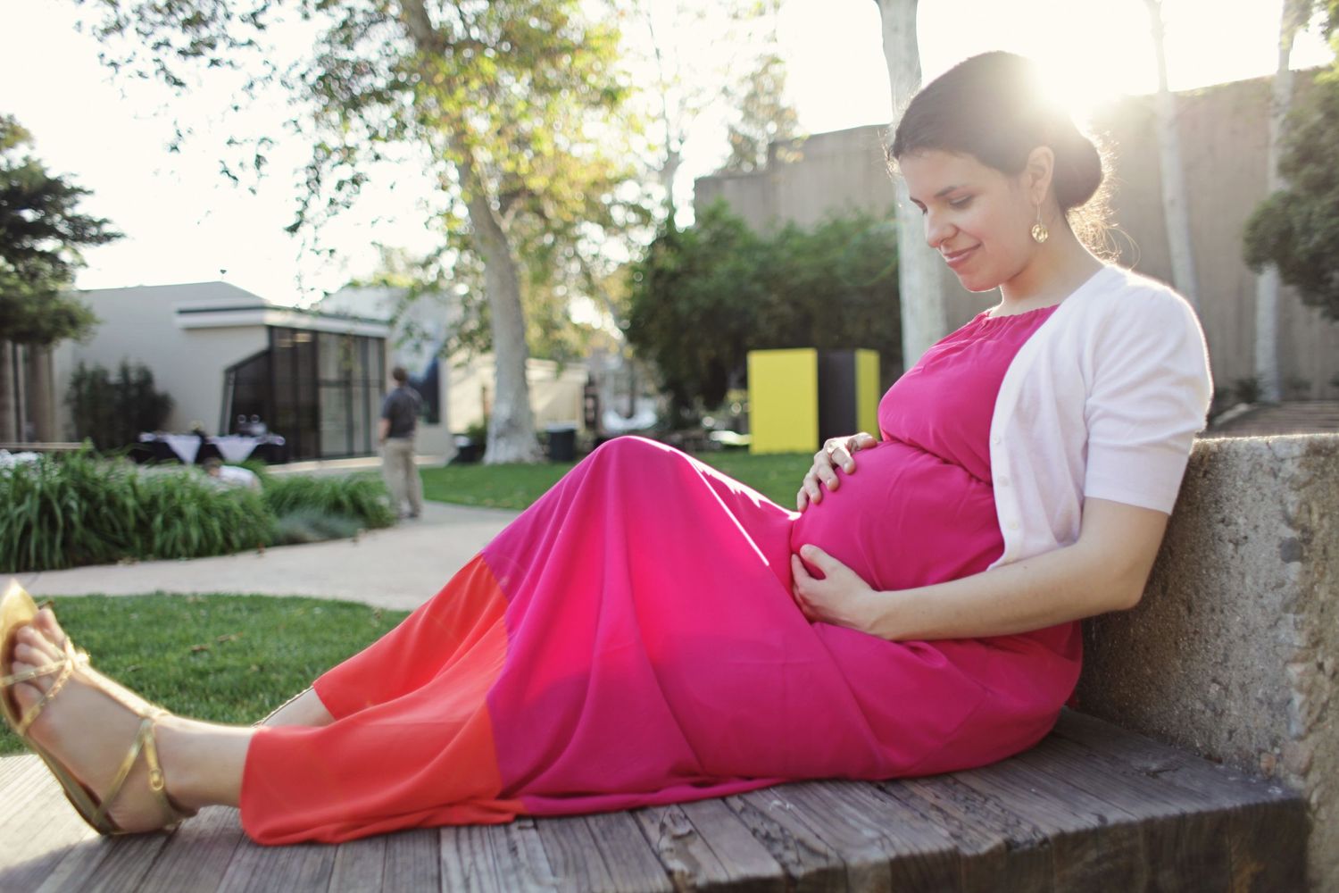 Expectant mother in red dress reading outdoors on bench.