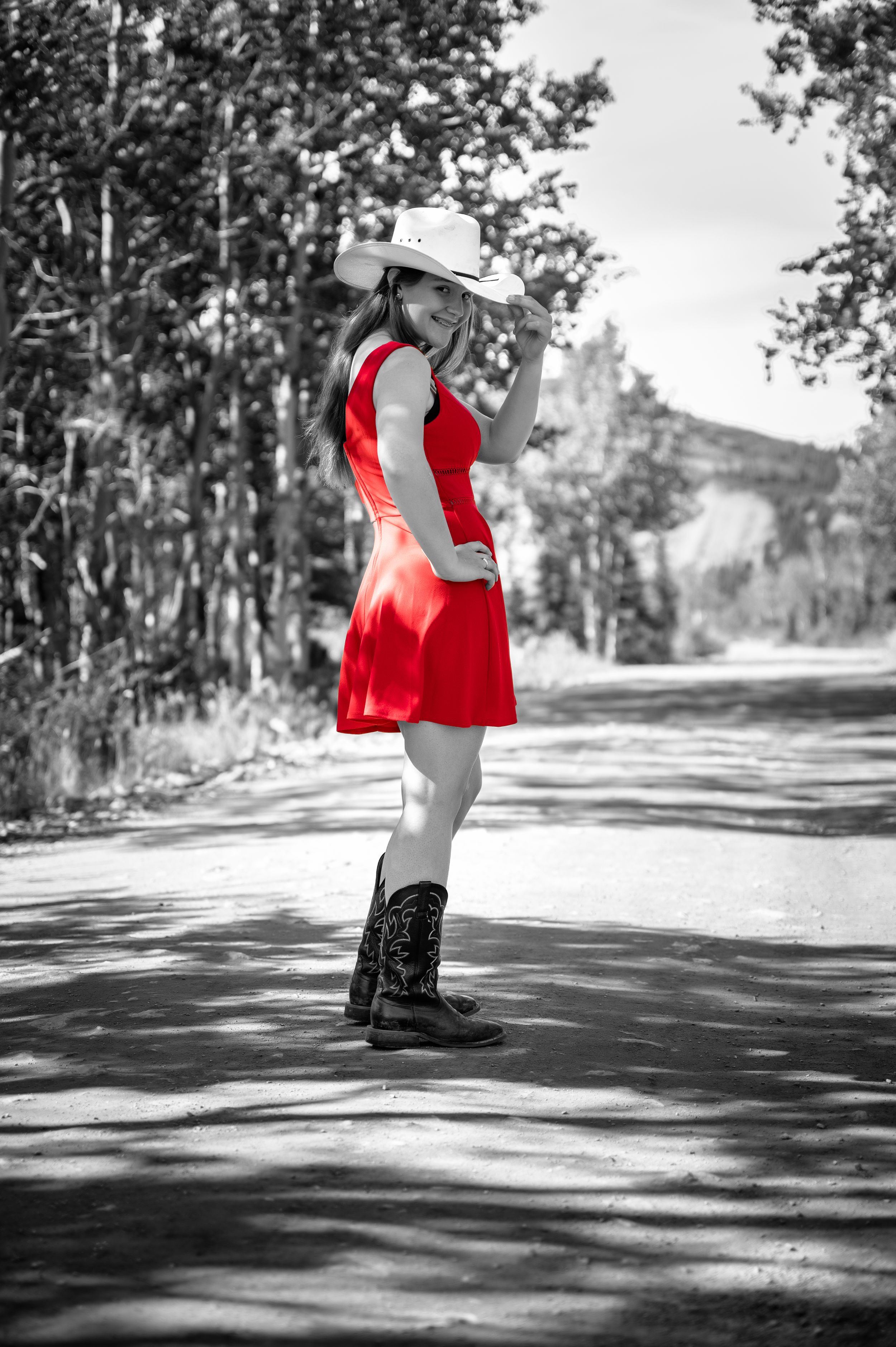 Figure in a red dress and cowboy boots poses on a rural mountain path in selective color photography.