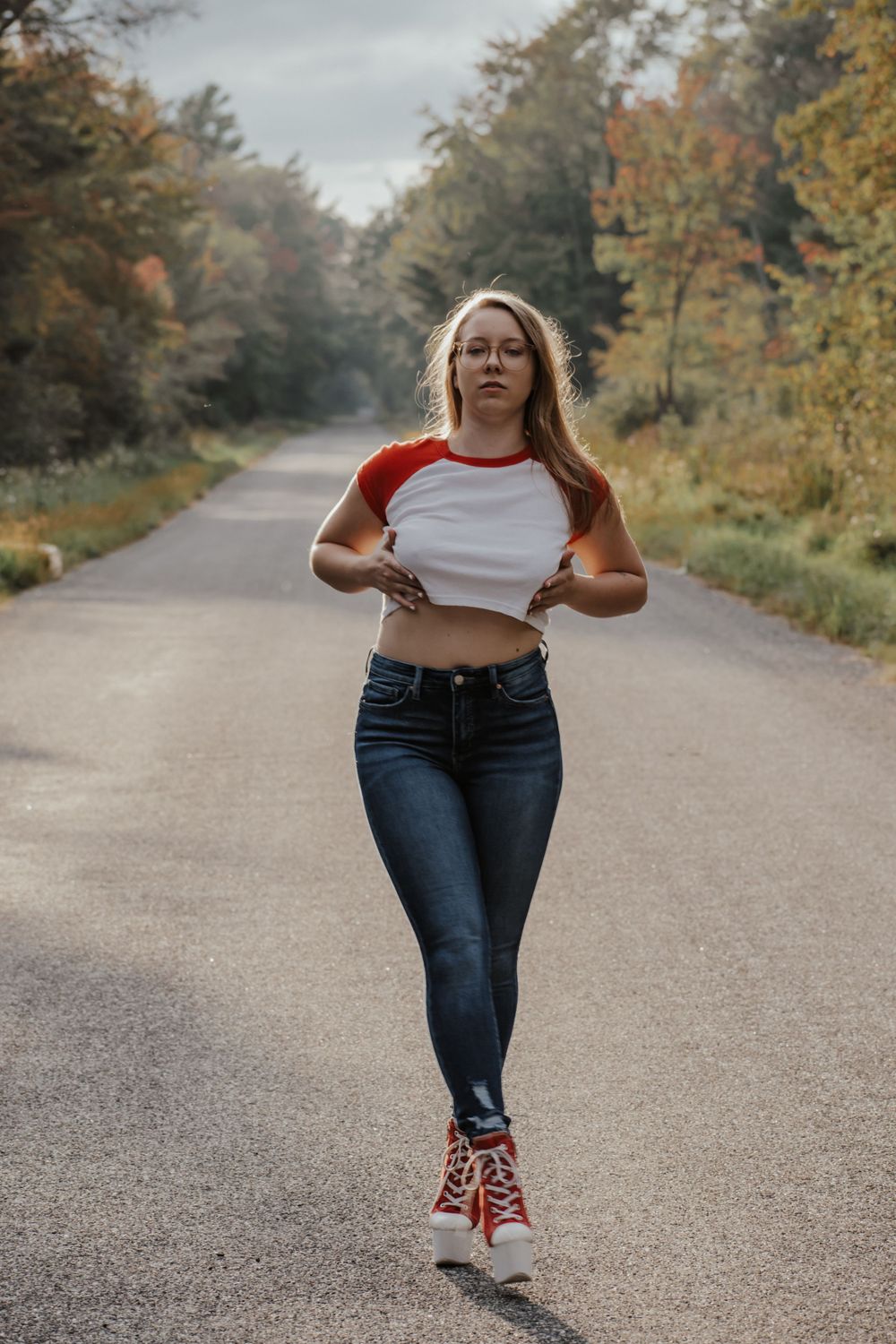 Athletic pose on rural road wearing crop top, jeans and red sneakers with fall foliage in background.