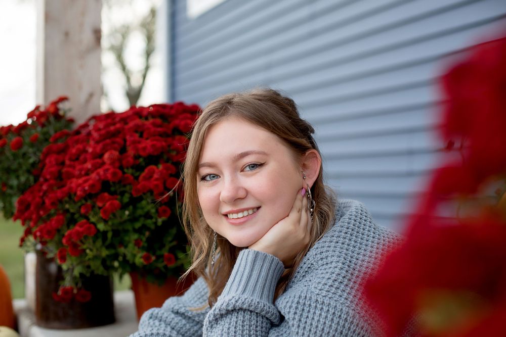 Portrait by red mums and blue siding wearing a cozy grey sweater and dangle earrings.