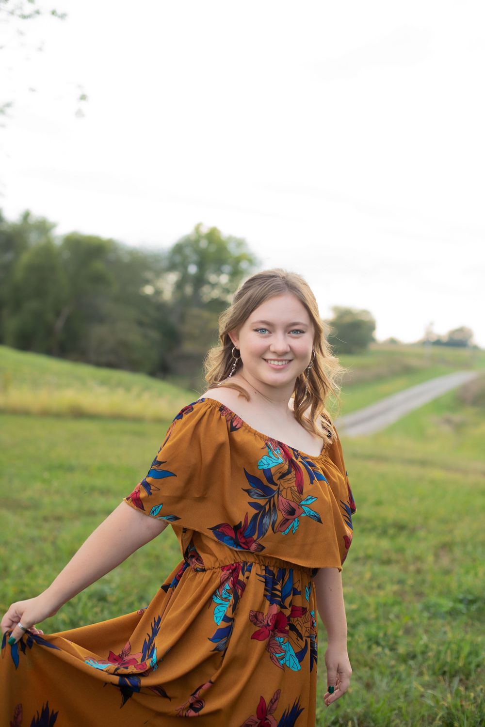 Standing in a rural field wearing a flowy rust-colored dress with blue floral pattern.
