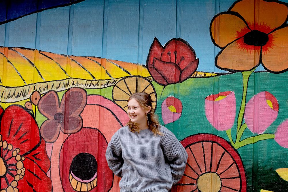 Person wearing gray knit sweater stands before vibrant floral mural artwork.