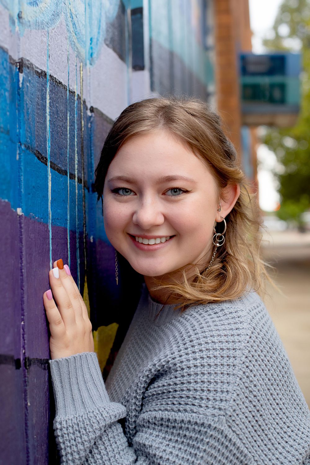 Person in gray sweater poses against colorful street art mural.