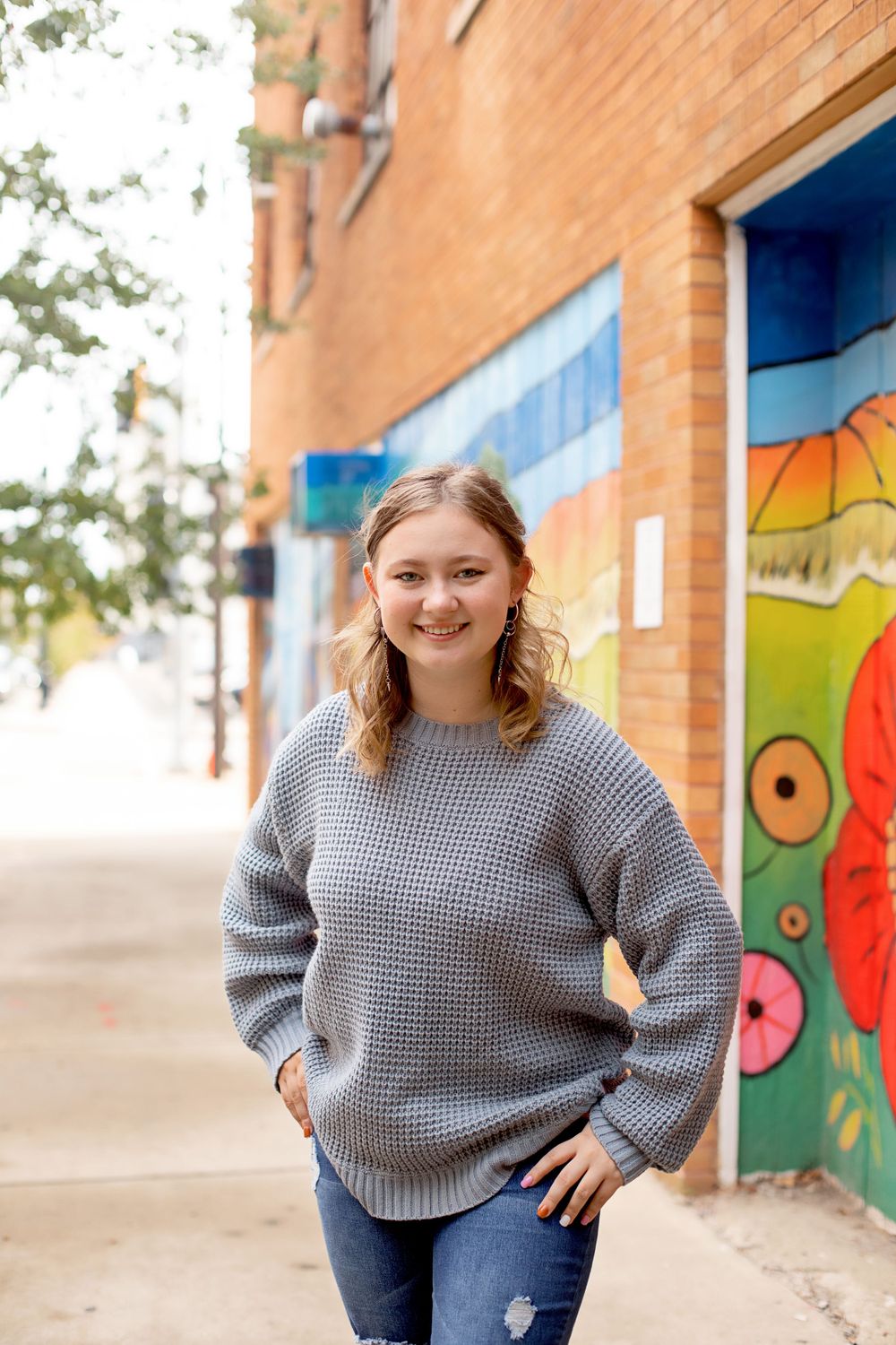 A casual portrait against a brick wall with colorful street art, wearing a grey knit sweater and jeans.