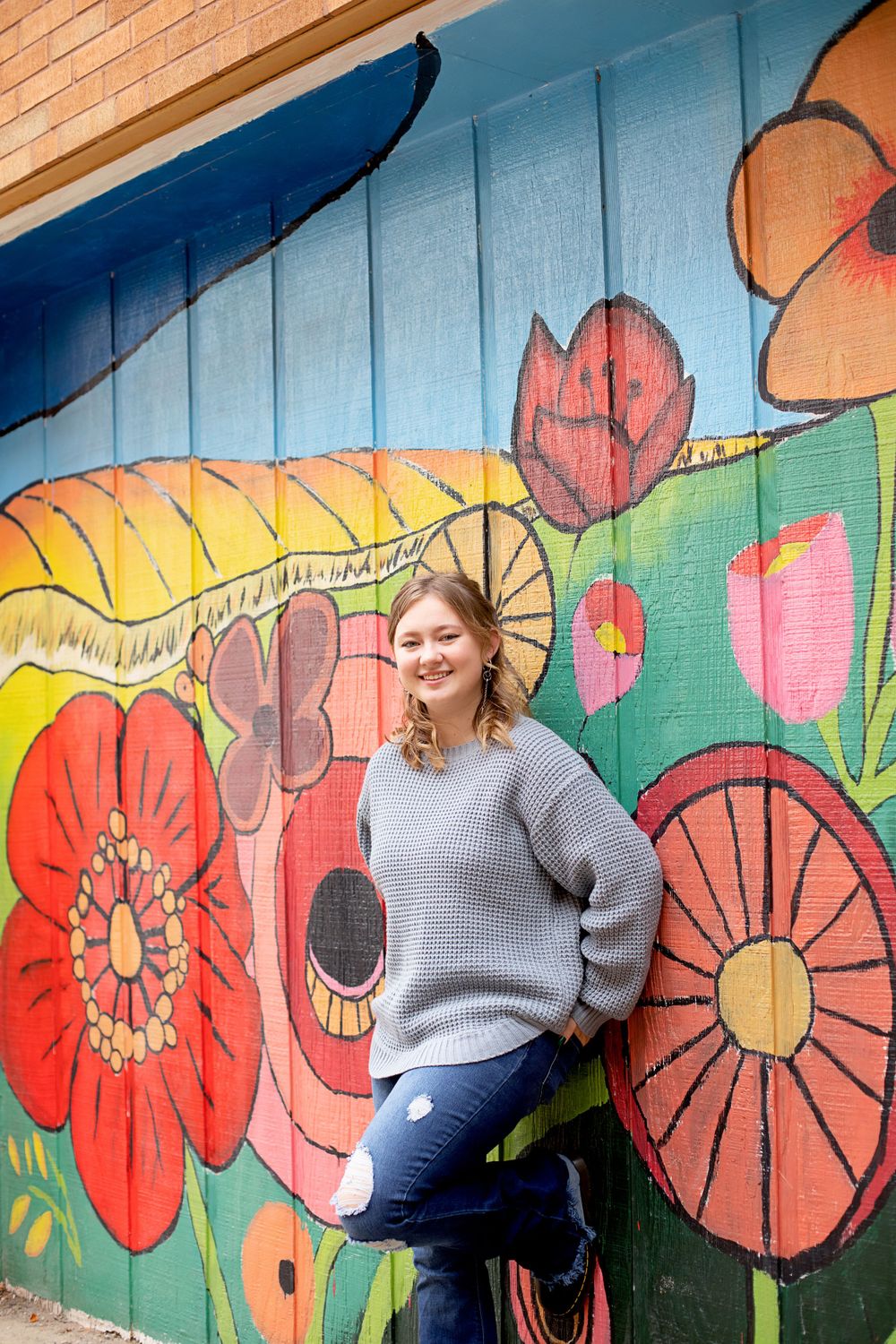 Person in cozy gray sweater poses against whimsical flower wall art.