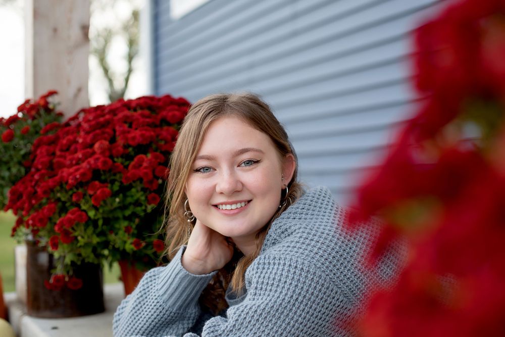 Portrait with red flowers and blue siding wearing a grey knit sweater.